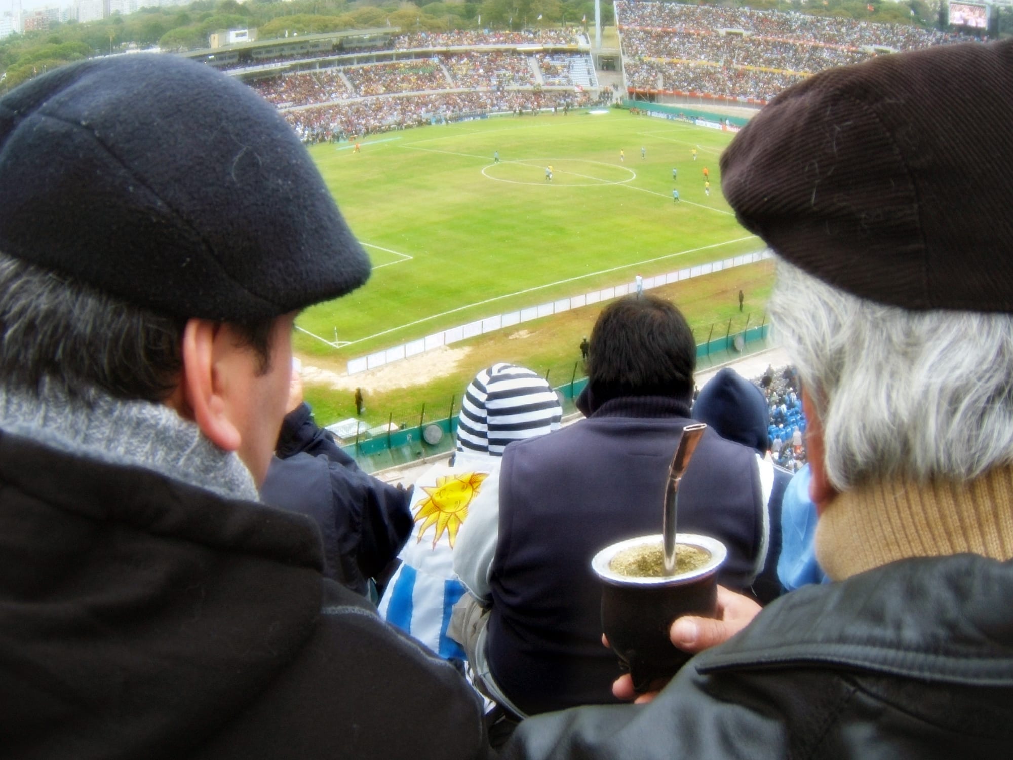 A man in a flat cap holds a dark mate gourd with a metal straw while watching a football match from high in a packed stadium, with a fan in a Uruguay flag below