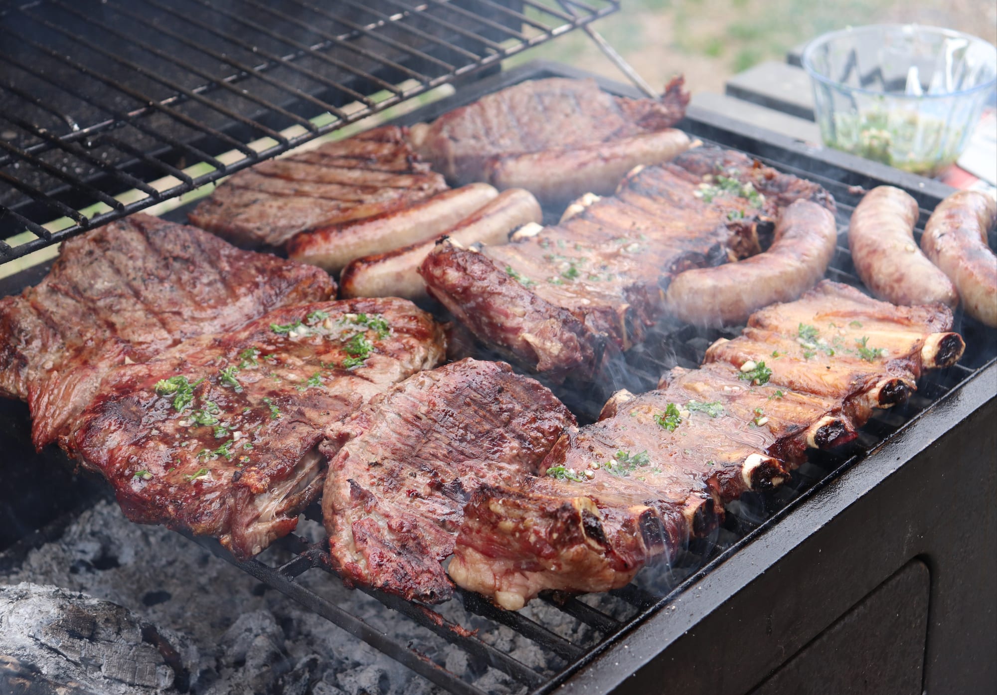Steaks, sausages and ribs grilling over open coals, garnished with chimichurri