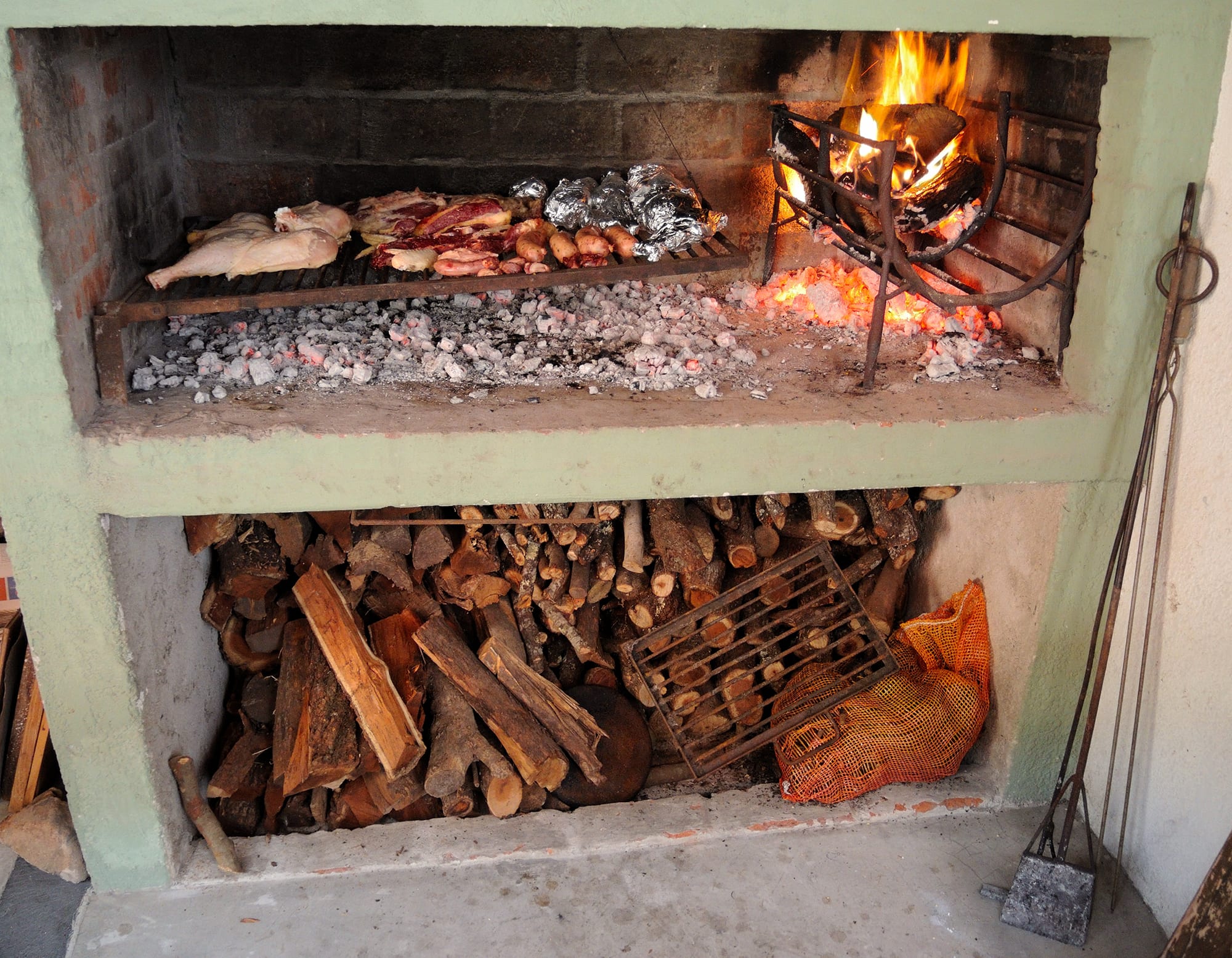 A built-in brick and concrete outdoor grill with meat and sausages cooking over hot coals, firewood stacked below