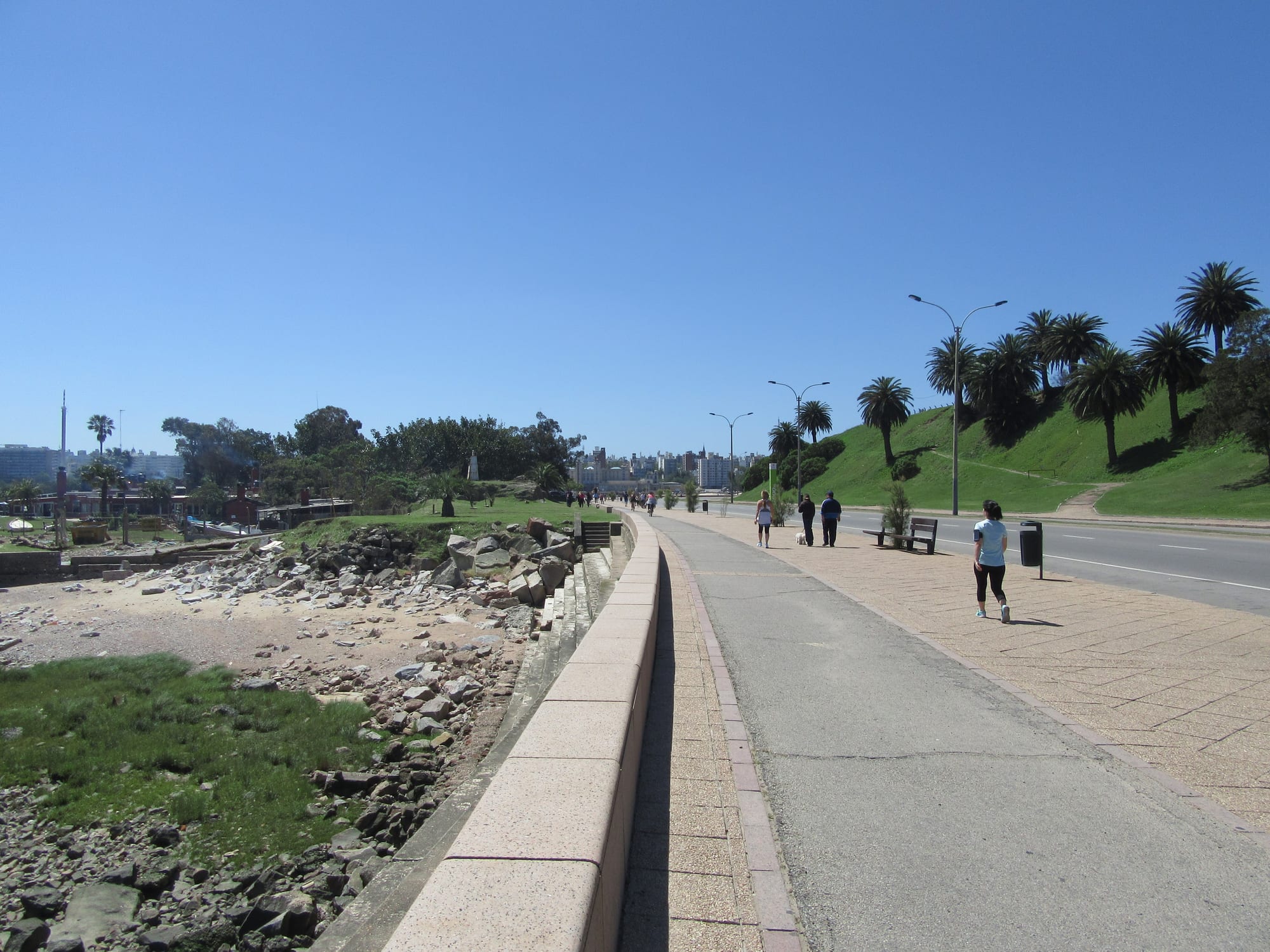 People walking and jogging along a wide coastal promenade lined with palm trees and street lamps beside the Río de la Plata in Montevideo