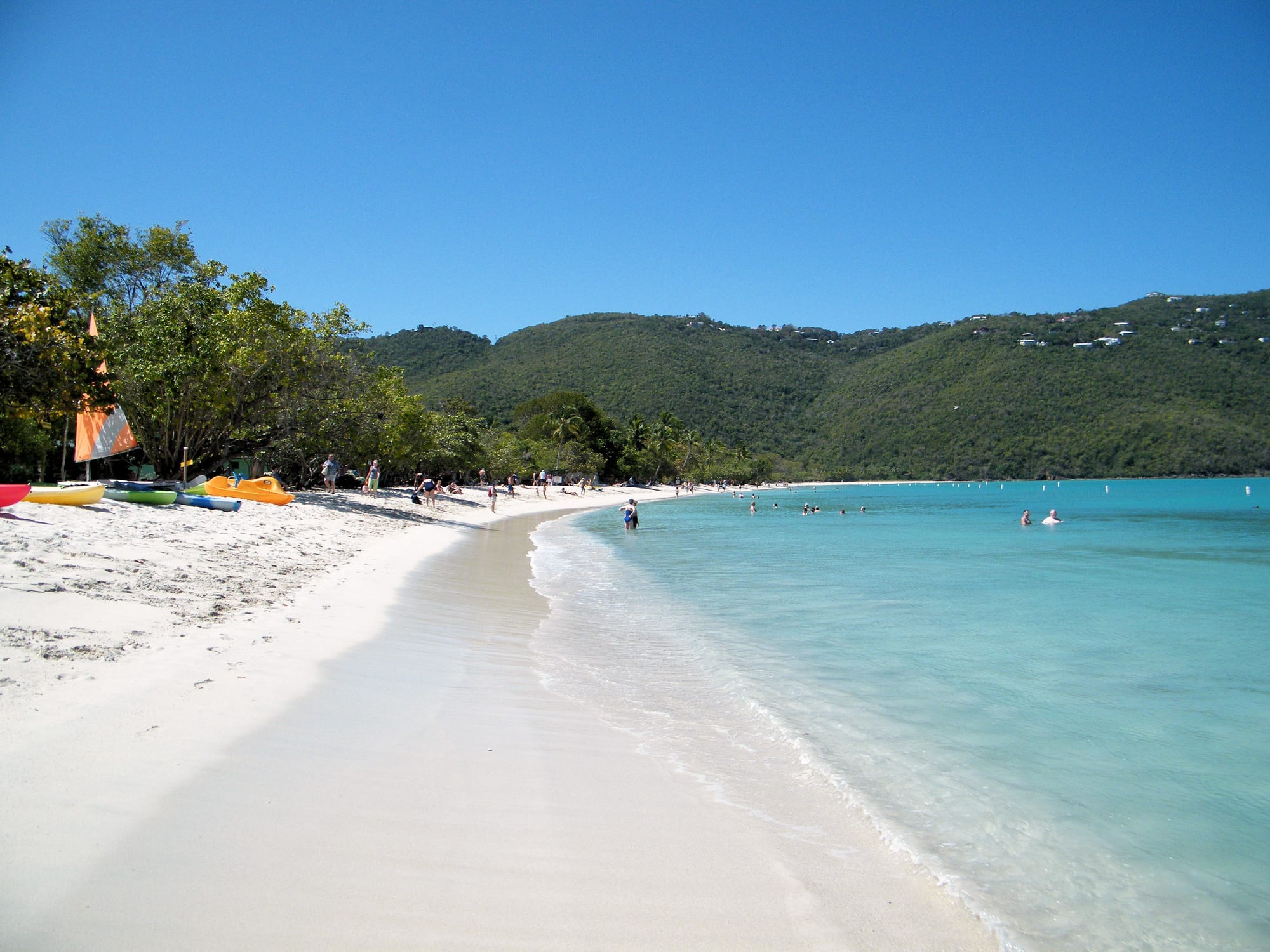 People swimming and relaxing on a white-sand beach with turquoise water and green hills at Magens Bay, St Thomas