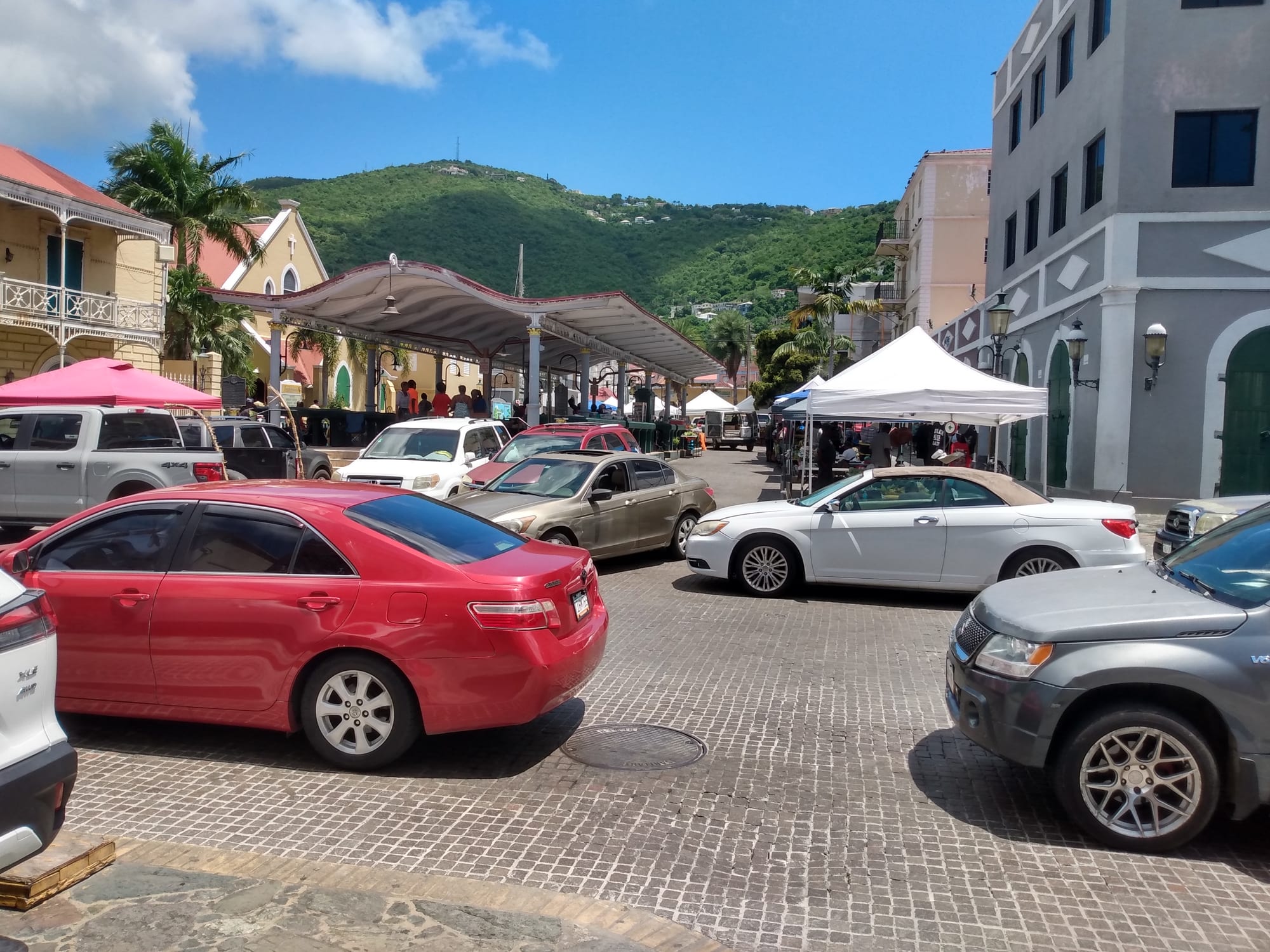 Cars parked around a market square with vendor tents, a church and green hills behind, in Charlotte Amalie