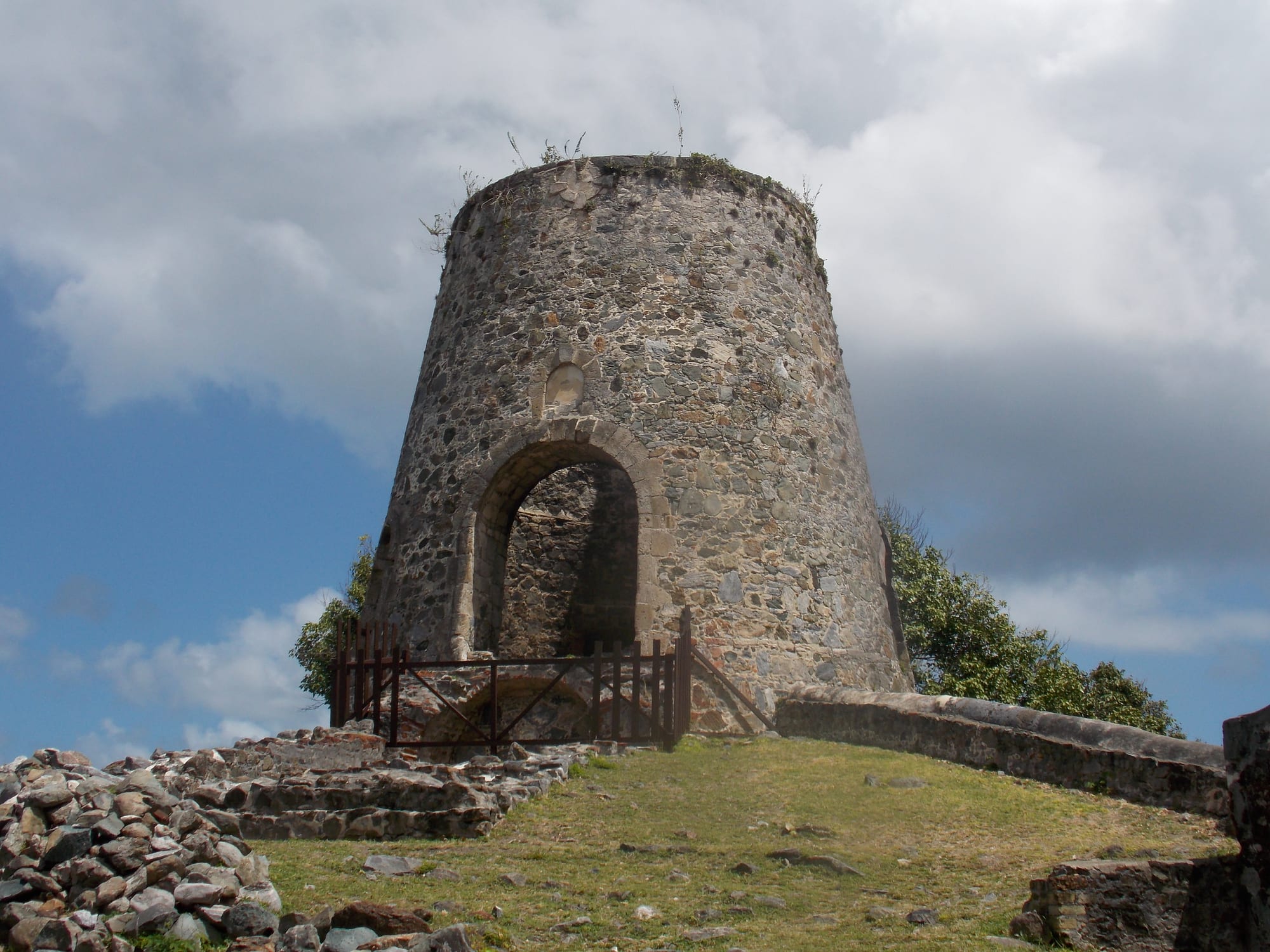 A round, tapering stone windmill tower with an arched doorway standing on a grassy rise beside low ruined walls, under a sky of heavy cloud and blue patches