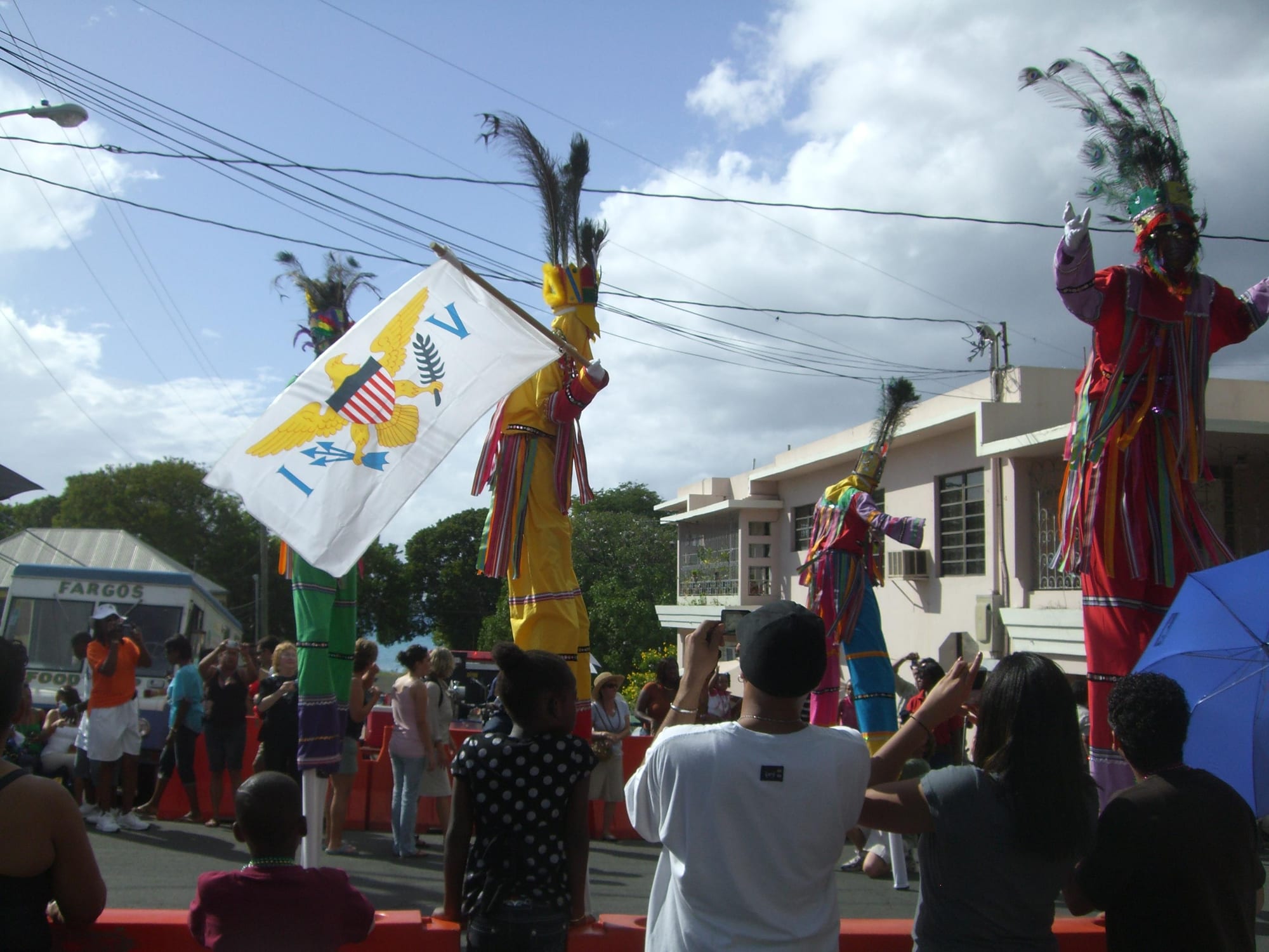 Four moko jumbies on tall stilts in brightly coloured costumes and feathered headdresses tower over a street crowd, one of them waving the flag of the US Virgin Islands