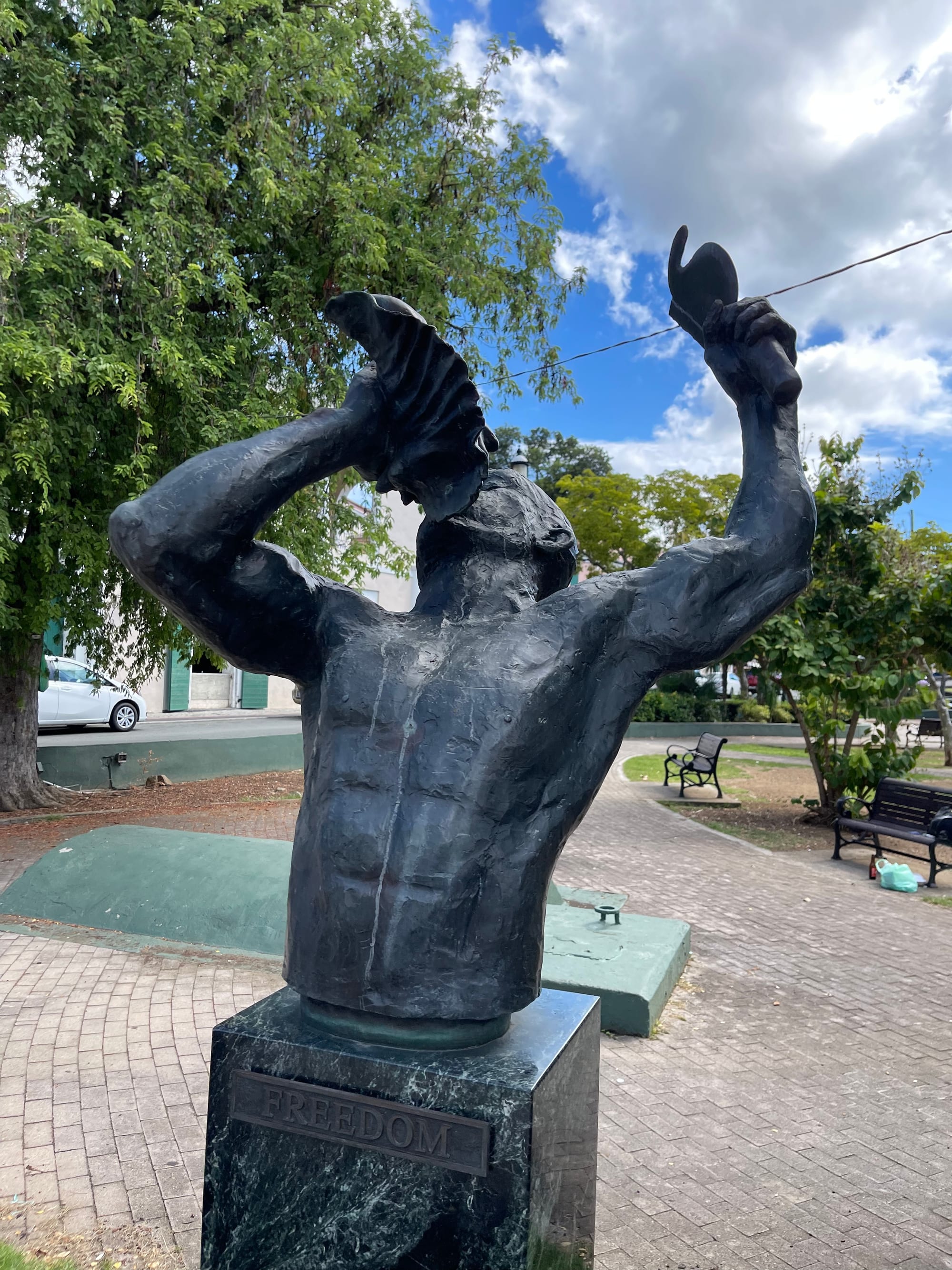A bronze bust of a man with both arms raised, blowing into a conch shell, on a dark marble plinth marked Freedom, in a paved garden with trees and benches