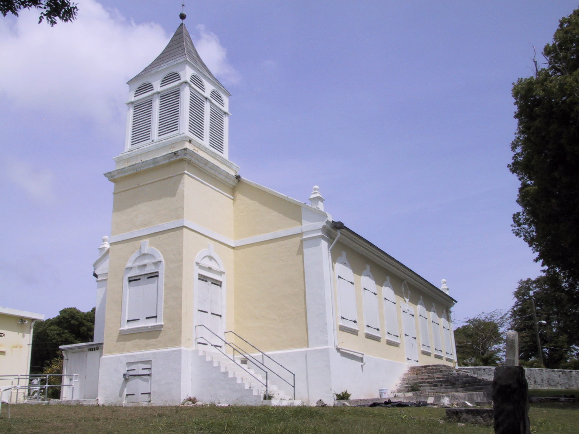 A pale yellow church with white trim, a louvred bell tower and tall shuttered windows, with stone steps and a lawn in front under a blue sky