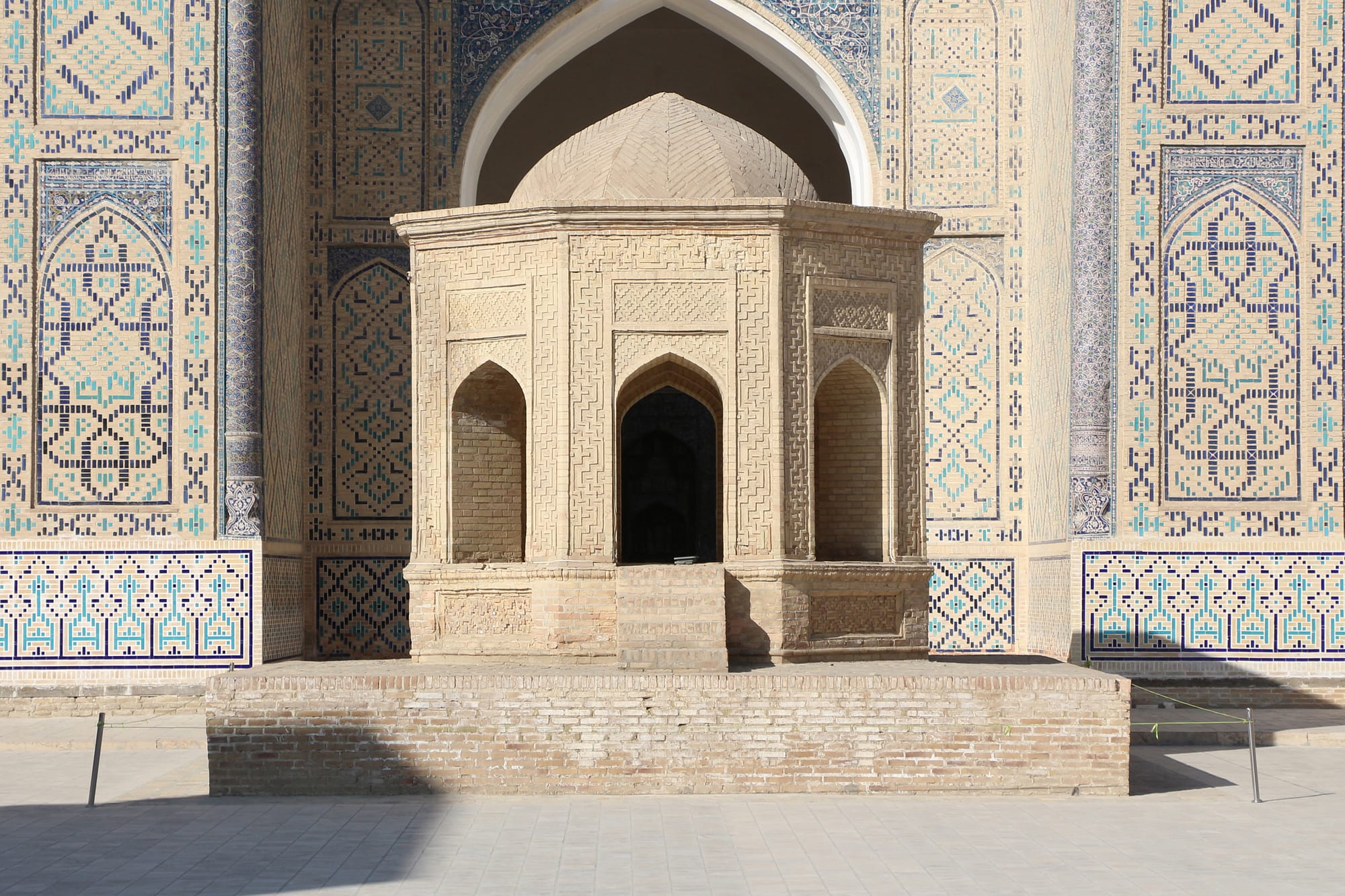 An octagonal brick pavilion with arched openings and a small dome stands before a tall arched portal decorated with blue and turquoise geometric tilework