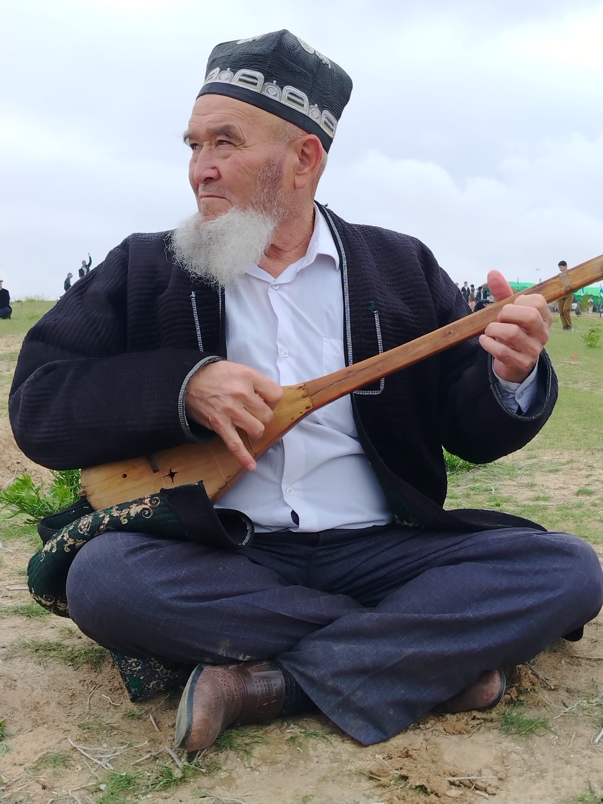 An elderly man with a white beard, an embroidered black skullcap and a quilted dark robe sits cross-legged on grass playing a long-necked wooden two-stringed instrument