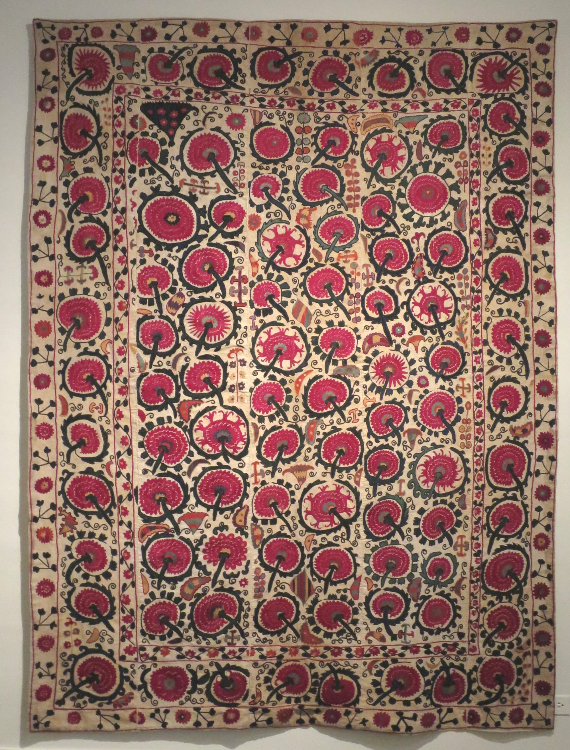A large embroidered textile with rows of big red and pink circular flower motifs and dark green stems on a cream ground, framed by a floral border