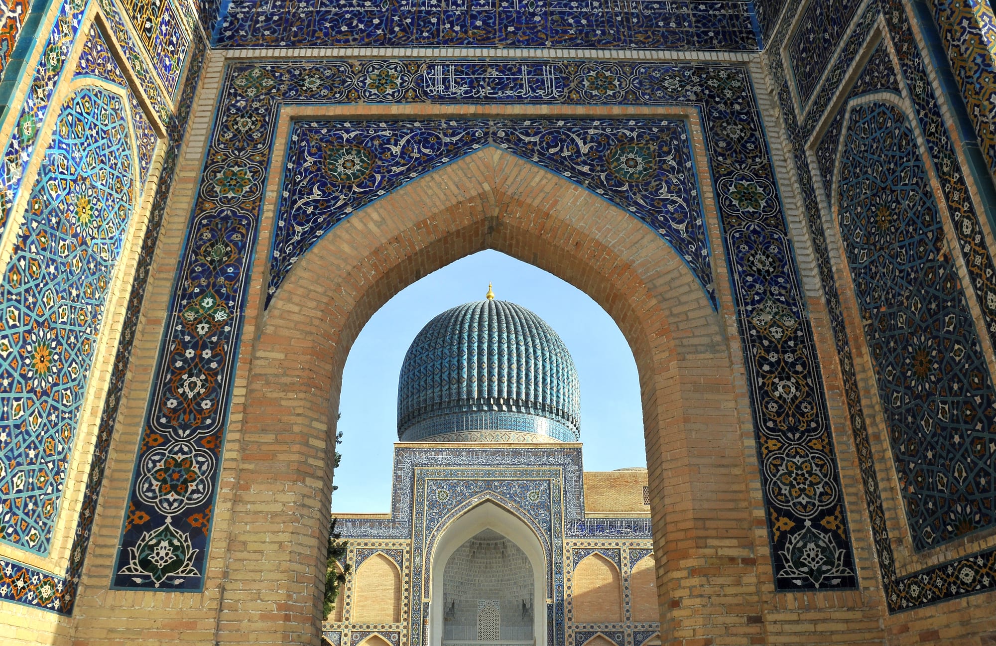 A ribbed turquoise dome seen through a tall pointed brick arch covered in blue, white and gold tilework