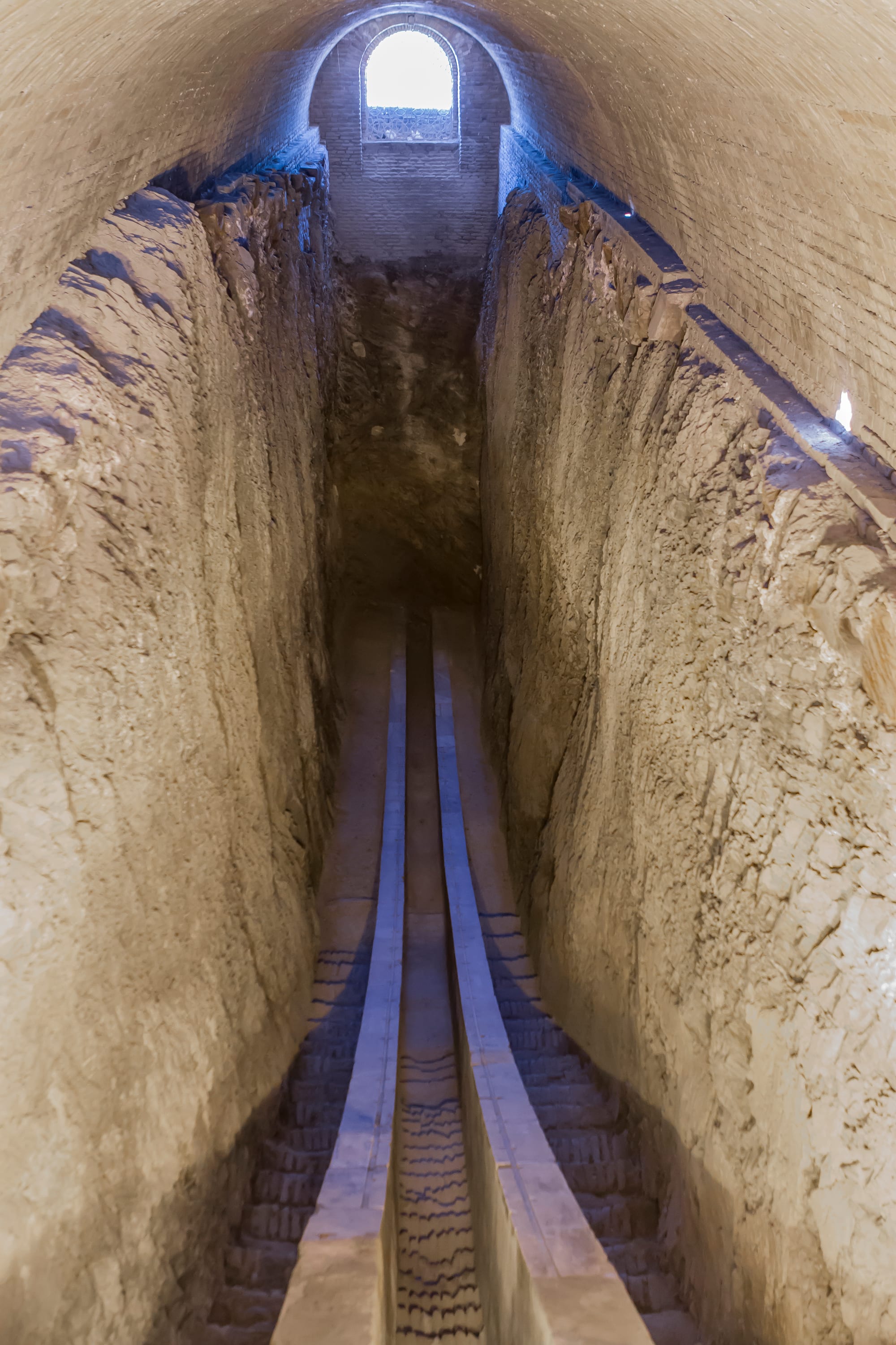 A long curved stone track with two parallel rails descending into a deep rock-cut trench under a brick vault, lit by a small arched window