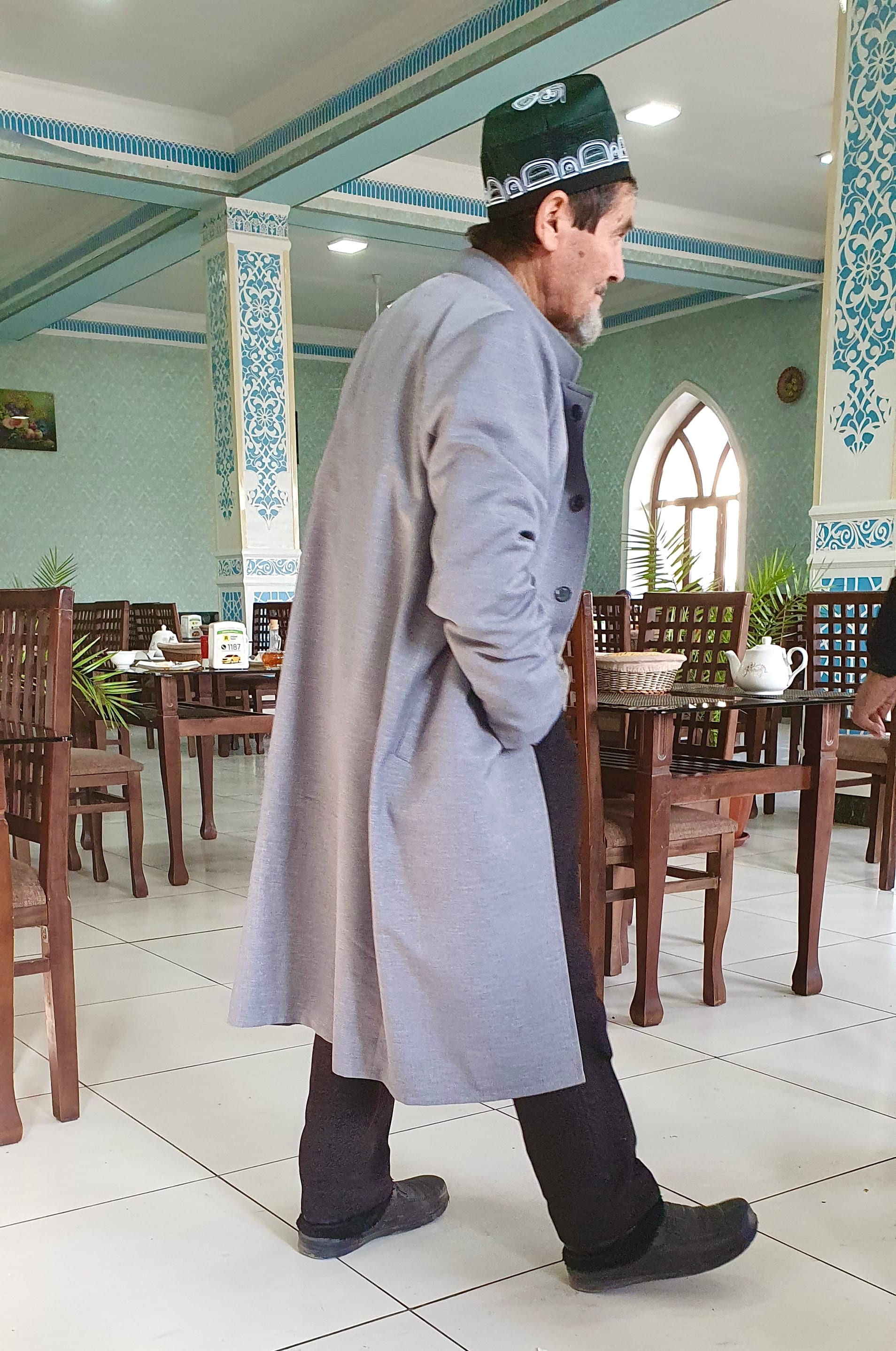 A man in traditional cap and coat walking through a decorated teahouse dining room set with tables and a teapot