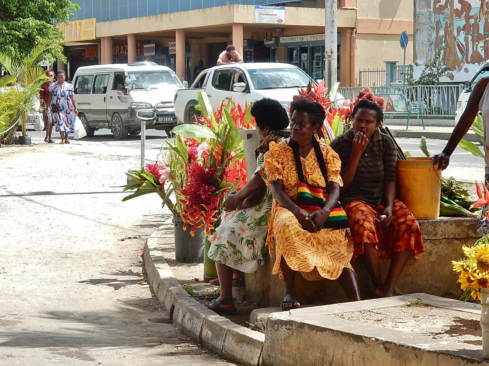 Ni-Vanuatu women in colourful island dresses sitting on a low wall beside buckets of tropical flowers on a Port Vila street