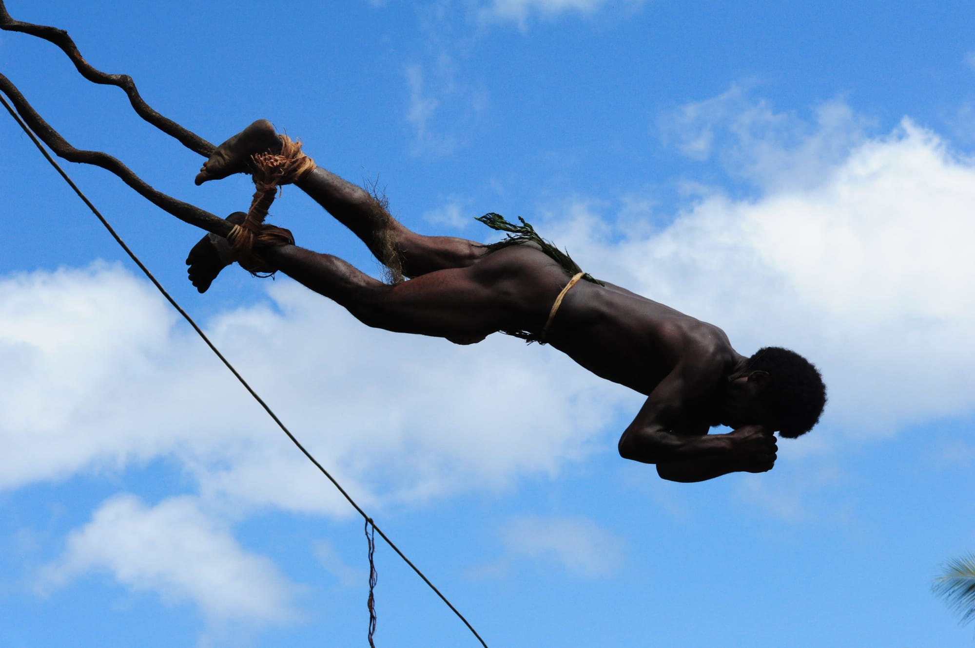 A man with vines tied around his ankles dives head-first through the air against a blue sky with white clouds