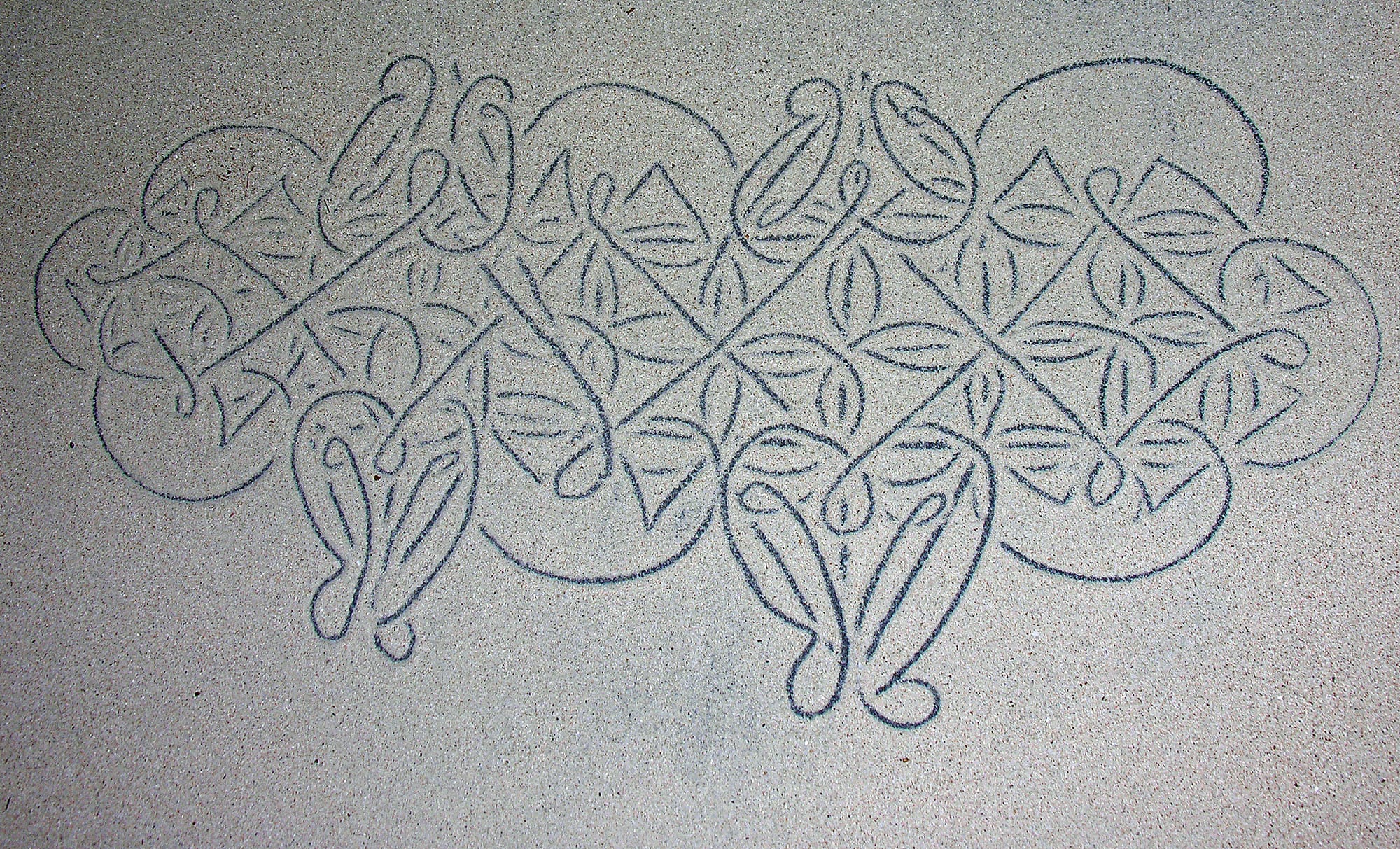 An intricate symmetrical pattern of looping lines, leaf shapes and diamonds traced in grey sand