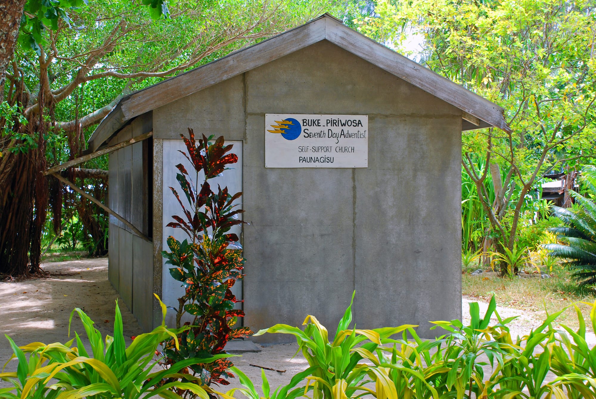 A small grey concrete church with a pitched roof and a painted sign reading Seventh Day Adventist Self-Support Church, surrounded by tropical plants and trees