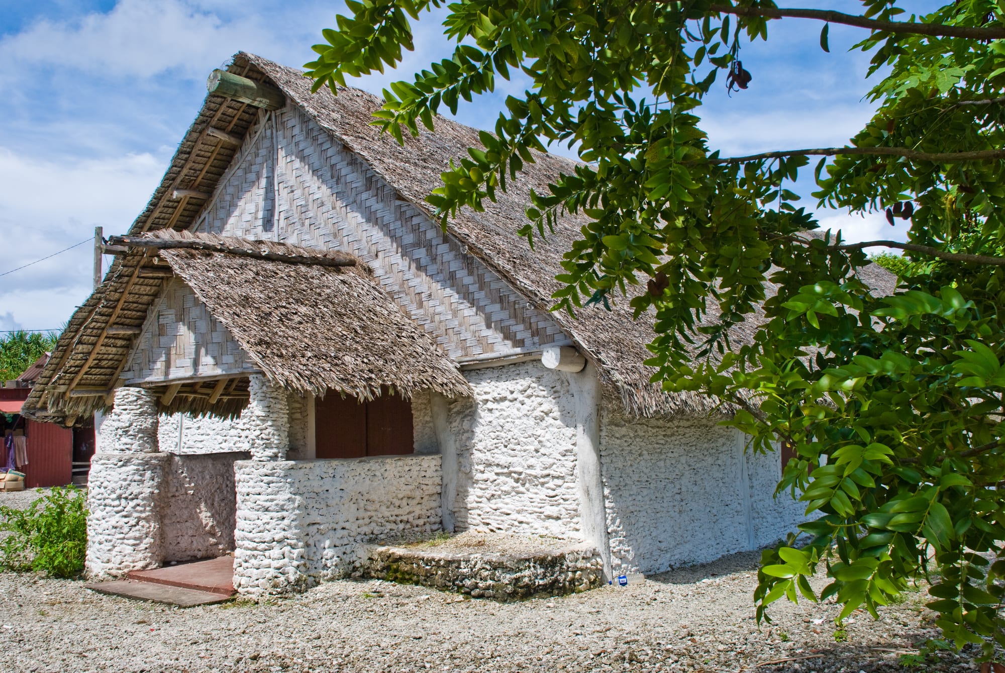 A long building with a thick thatched roof, woven bamboo gable walls and whitewashed stone walls, with a small porch, beneath a leafy tree
