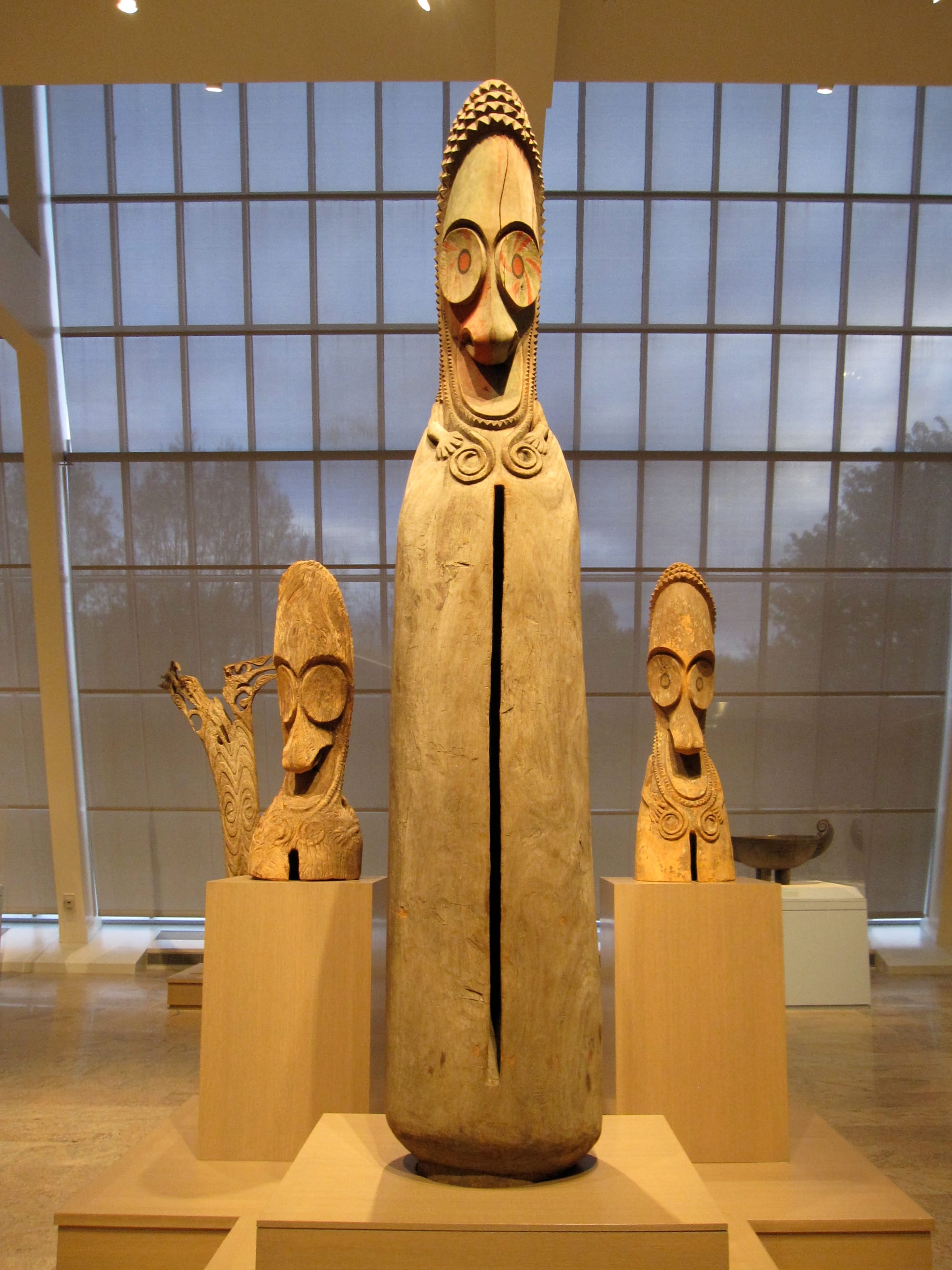 Tall carved wooden slit drums with long faces and lozenge-shaped eyes, standing upright side by side