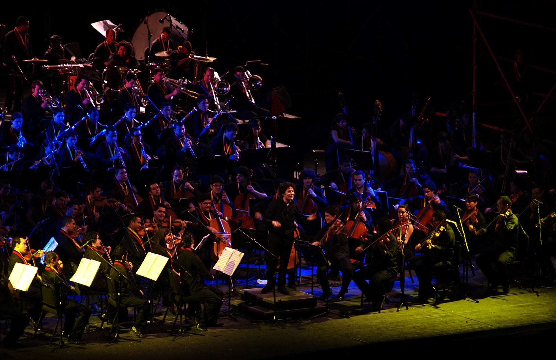 A large orchestra in dark clothes and orange scarves plays on a dimly lit stage, with a conductor at the centre and brass and percussion rising behind