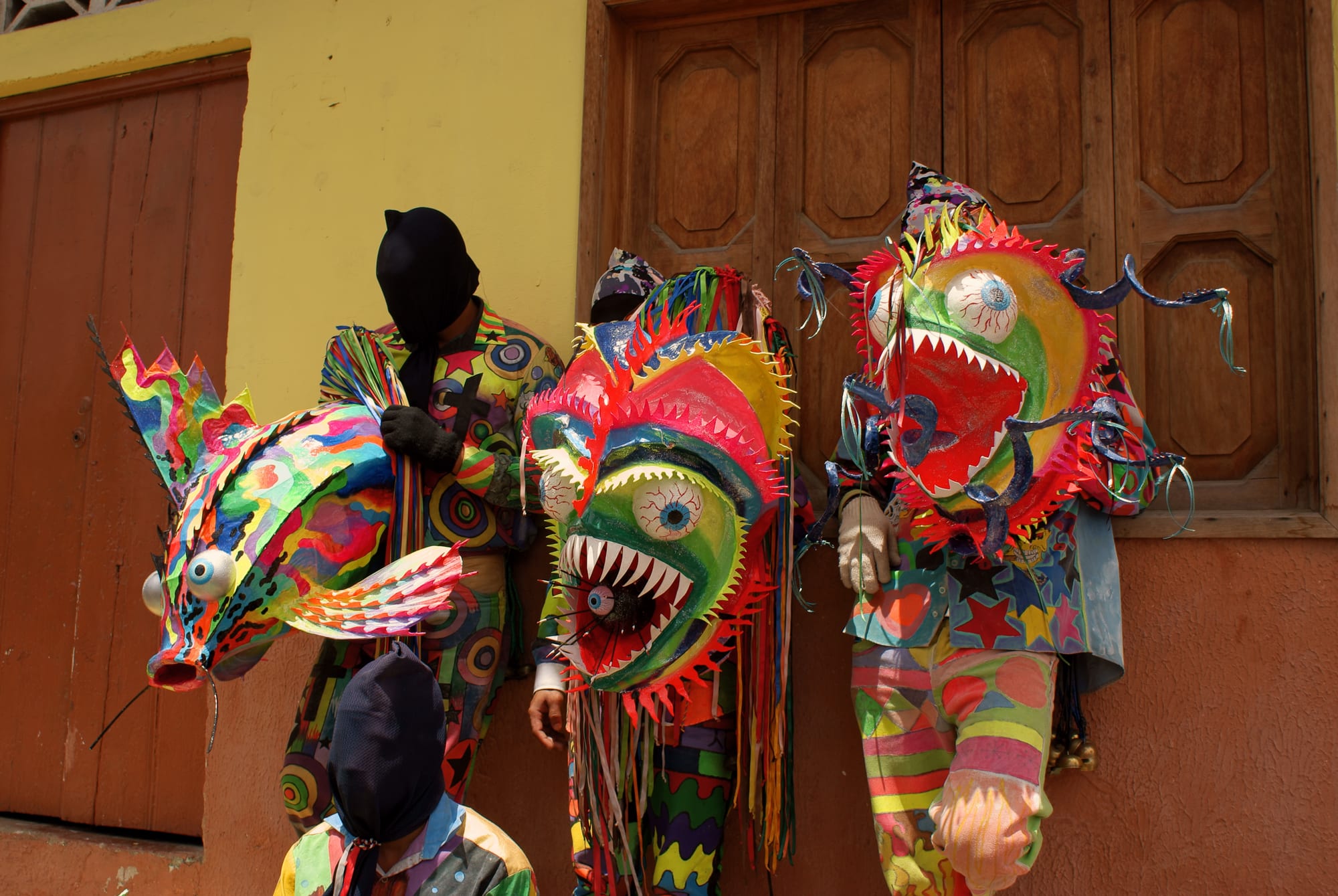 Three masked dancers in brightly patterned costumes stand against an orange wall and a wooden door, holding huge painted masks with bulging eyes and rows of sharp teeth