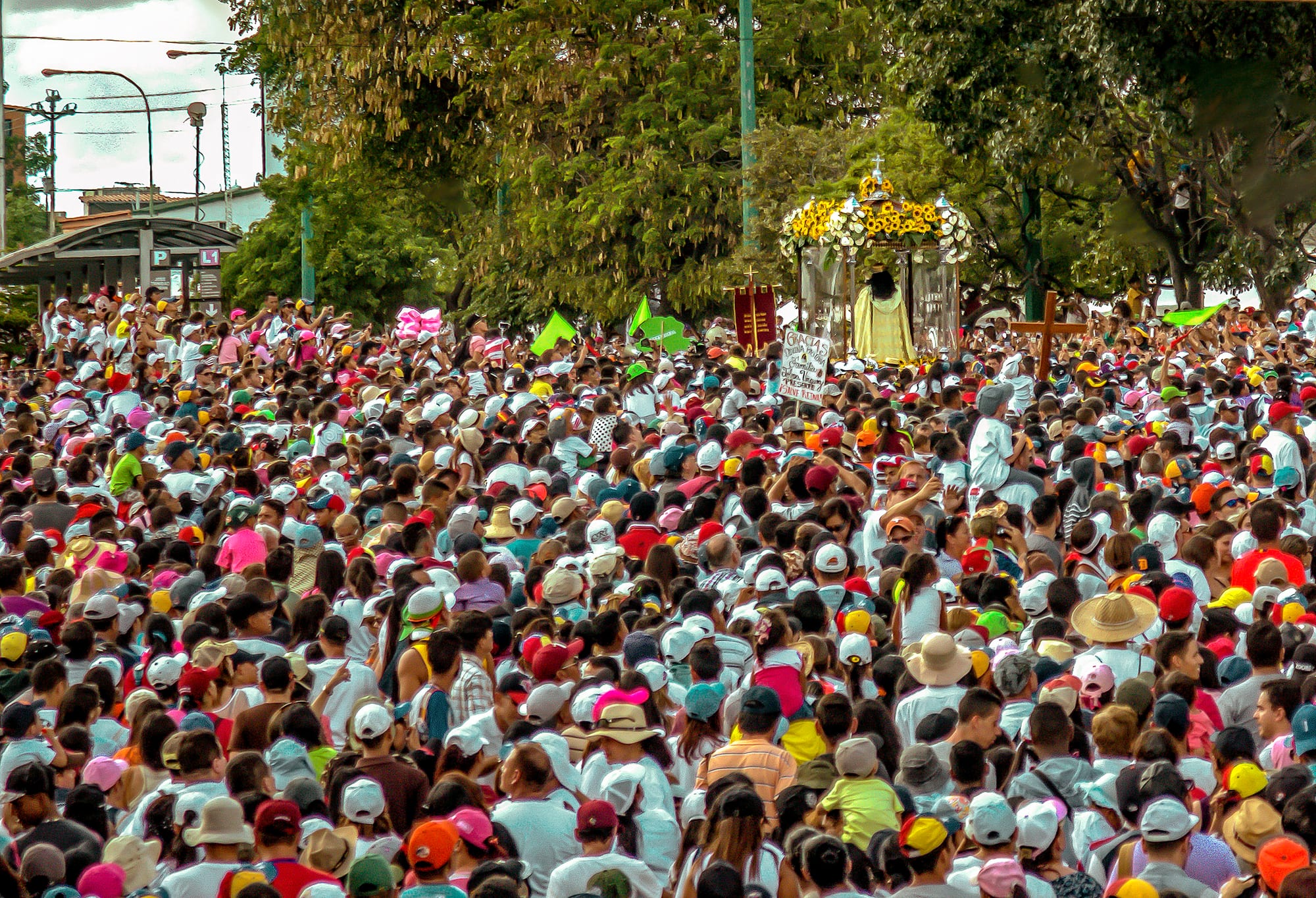 A dense crowd of people, many in white shirts and caps, fills a wide street around a canopied platform carrying a statue decorated with yellow flowers