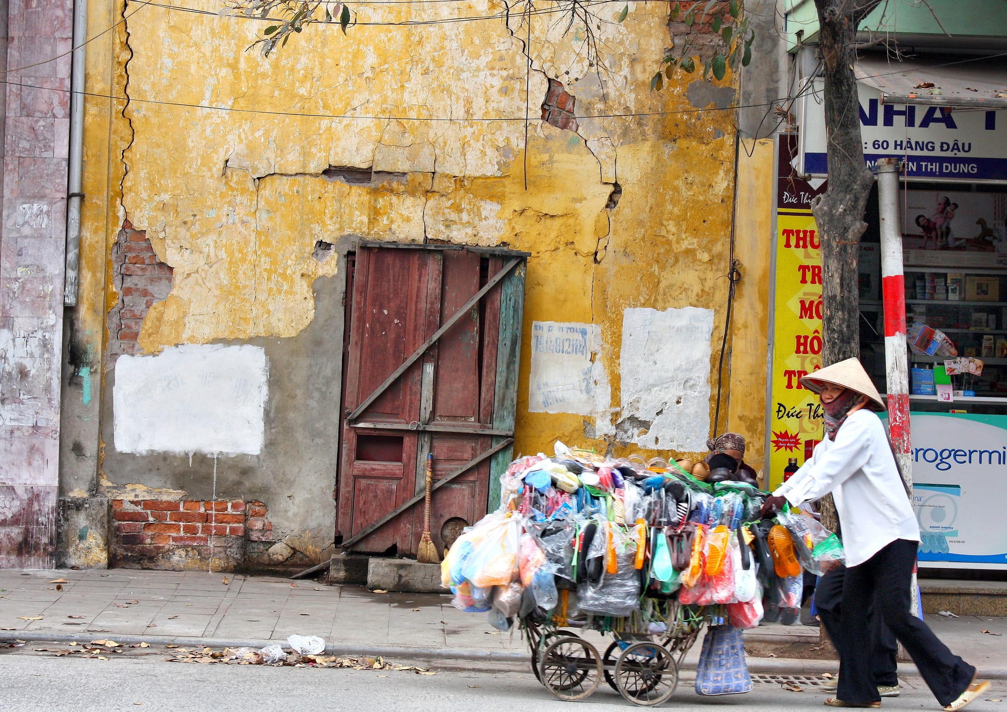 A street vendor in a conical hat pushing a cart piled with sandals and household goods past a weathered yellow wall in Hanoi
