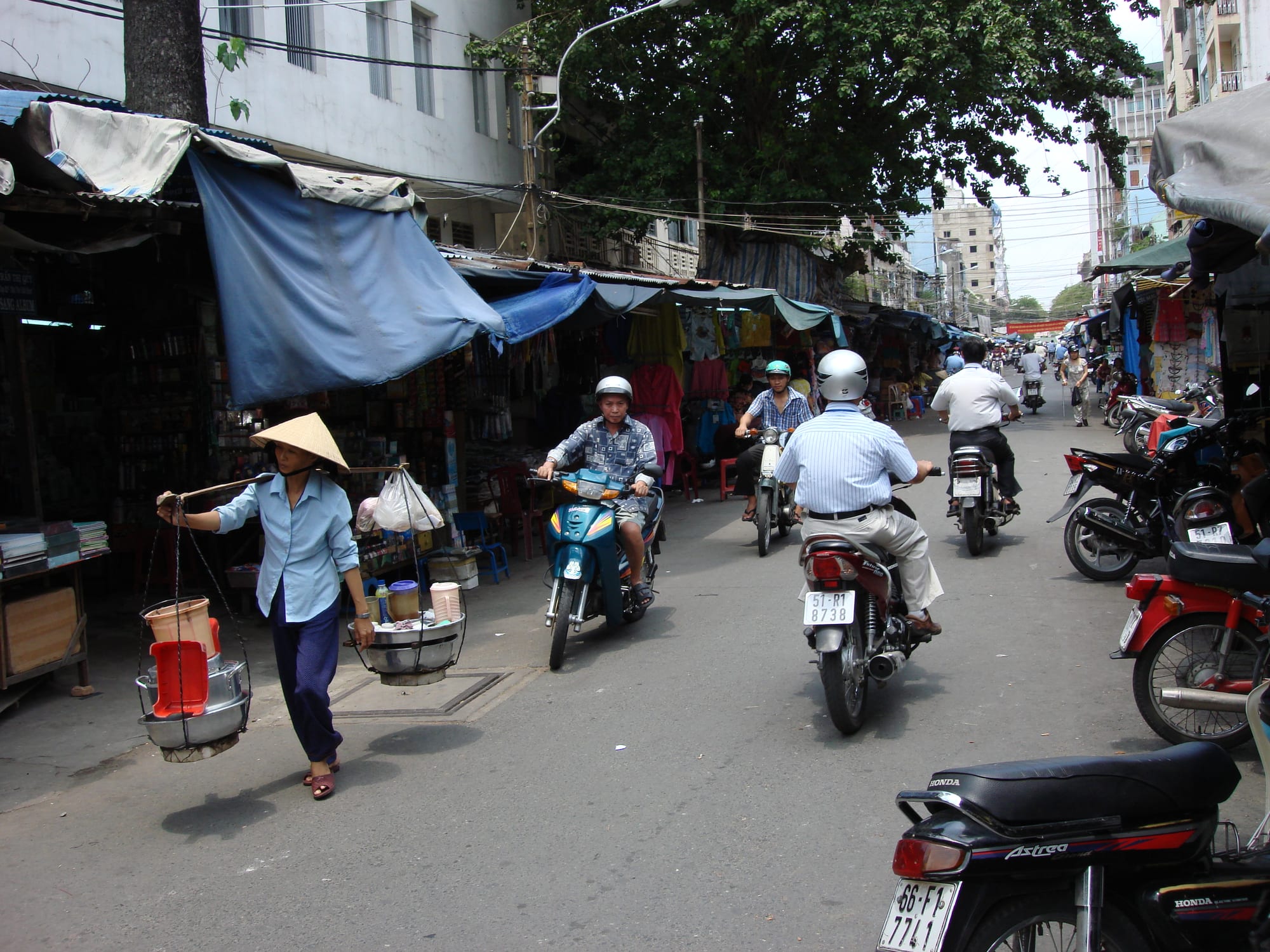 A narrow Vietnamese street lined with market stalls under awnings, motorbikes weaving past a street vendor carrying goods on a shoulder pole