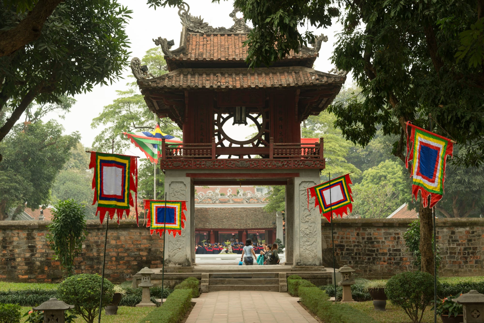A red wooden two-storey pavilion with a tiled roof and round windows standing on four white stone pillars, flanked by colourful banners, old brick walls and trees