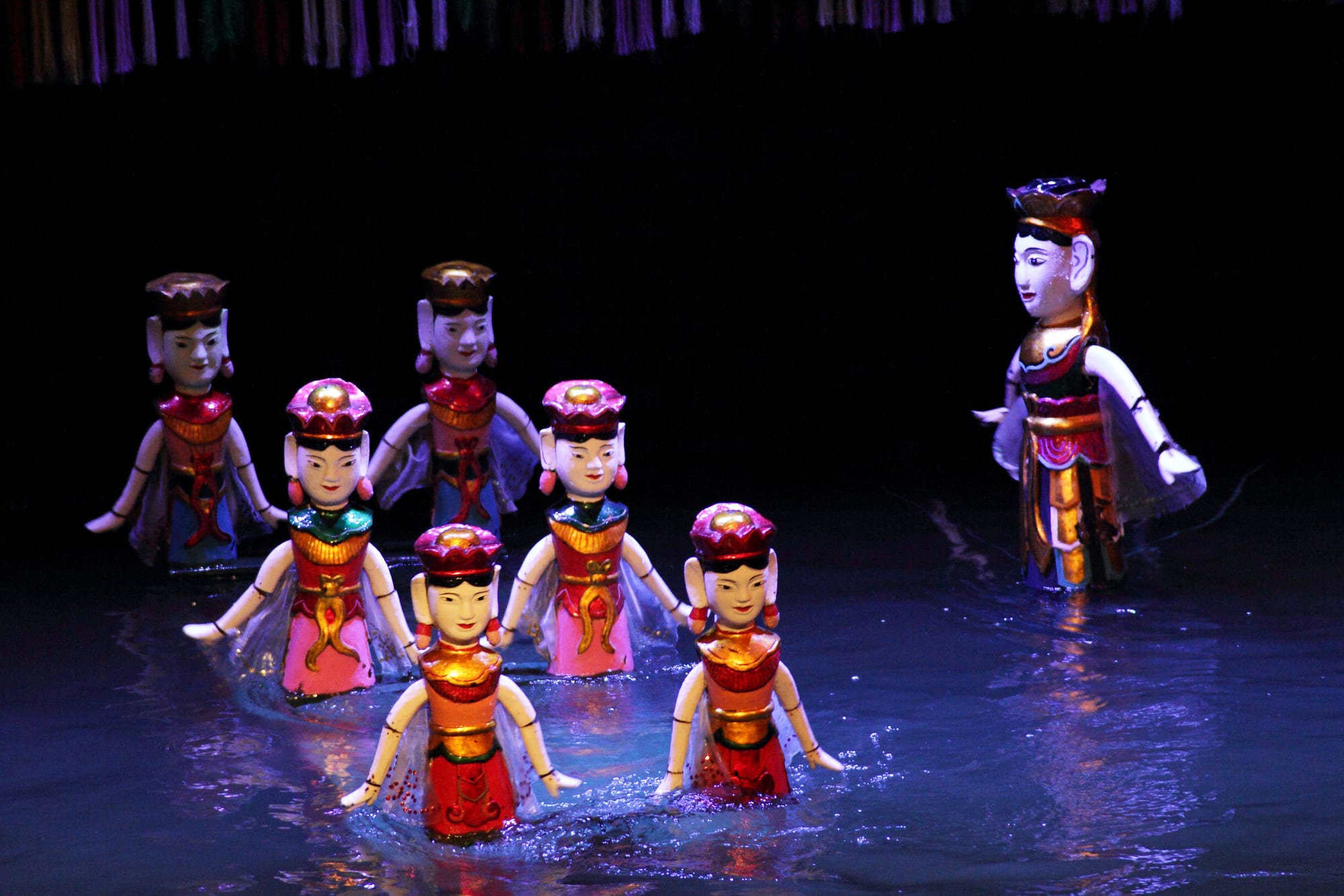 Brightly painted wooden puppets of dancing women in red and gold costumes glide across dark blue water, with a larger puppet figure standing to one side