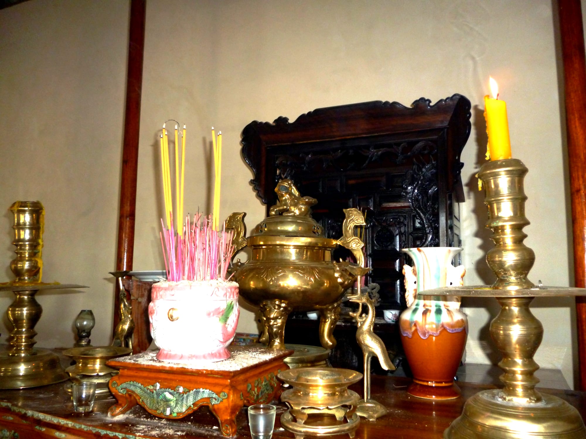 An ancestral altar with a pot of pink and yellow incense sticks, a brass incense burner, tall brass candlesticks, a lit yellow candle and a carved dark wooden shrine behind