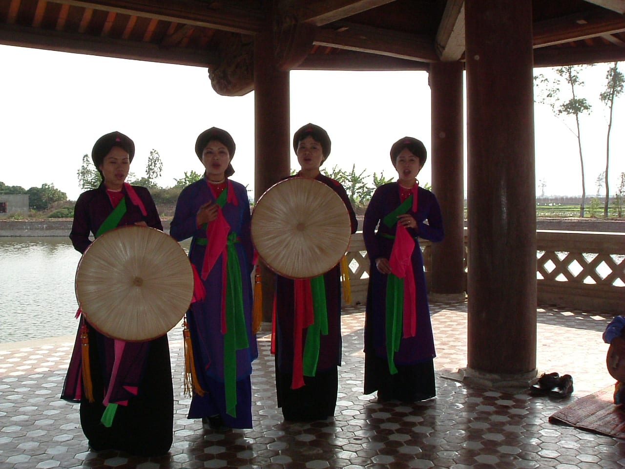 Four women in purple and dark tunics with pink and green sashes sing in a wooden pavilion beside a pond, two of them holding large round flat hats