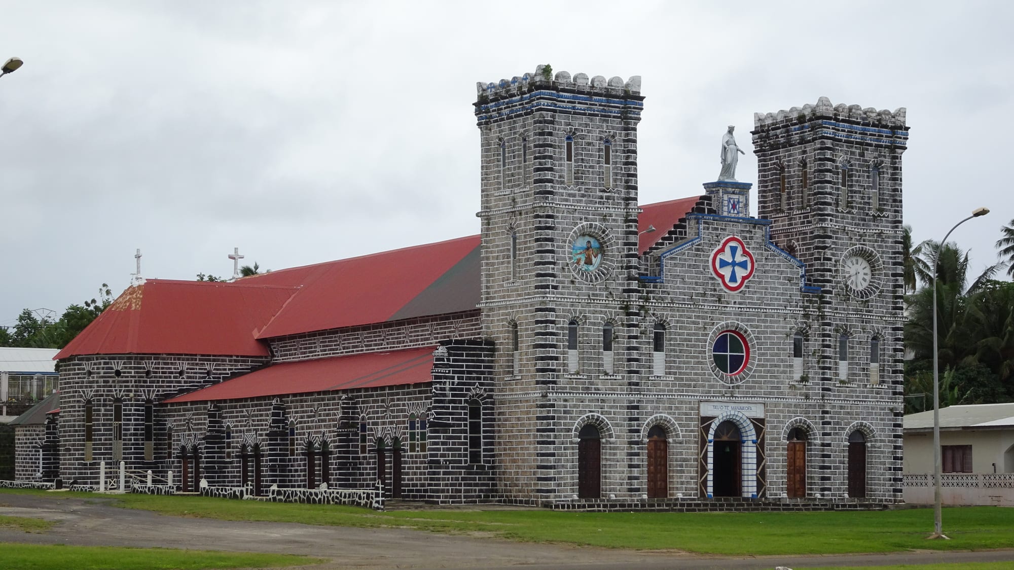 The black-and-white stone Cathedral of Our Lady of the Assumption in Mata-Utu, with twin bell towers and a red roof under a cloudy sky