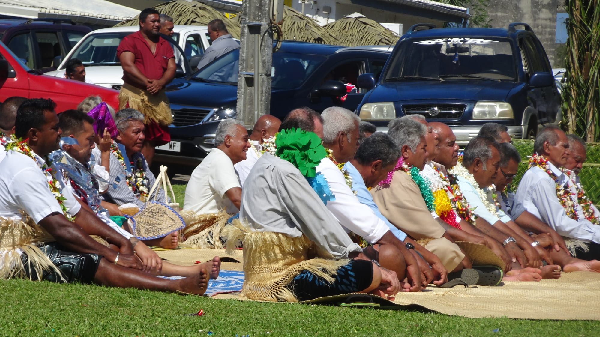 A row of men wearing flower garlands and woven waist mats sit cross-legged on large mats spread on the grass, facing forward, during an outdoor ceremony