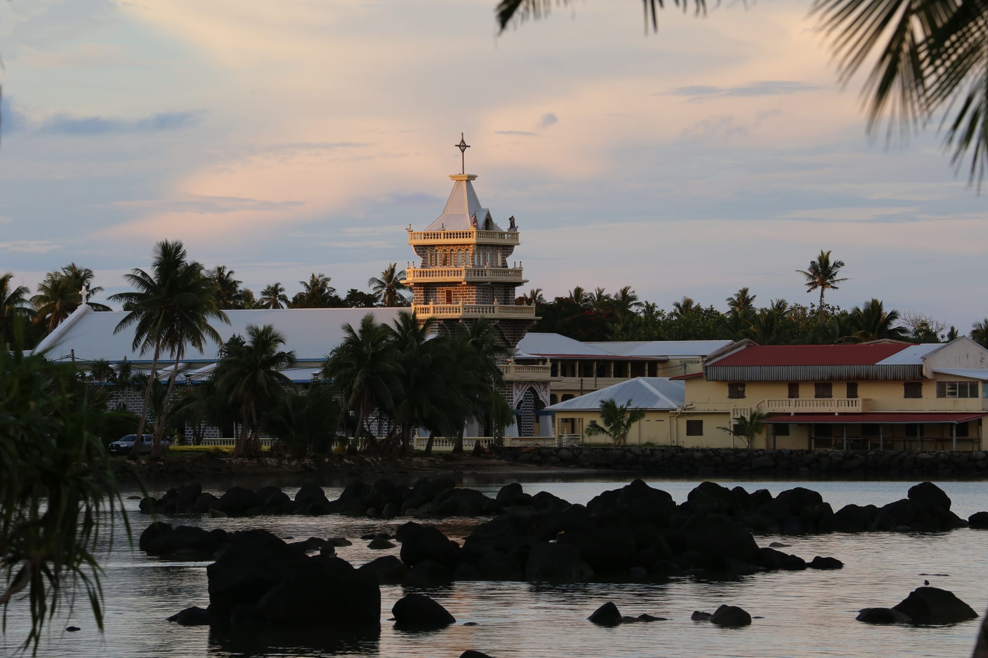 A tall pale church tower topped with a cross rises behind palm trees at dusk, reflected in calm water scattered with dark volcanic rocks