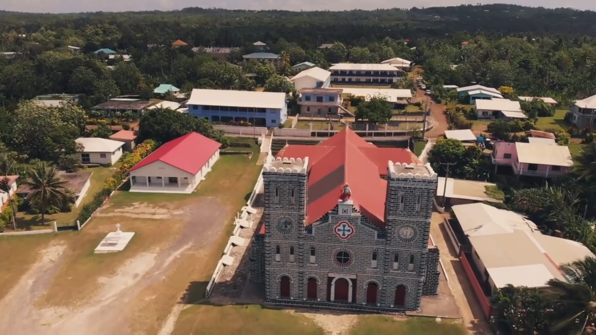 A large twin-towered stone Catholic cathedral surrounded by palms and low houses beside a lagoon at Mata-Utu