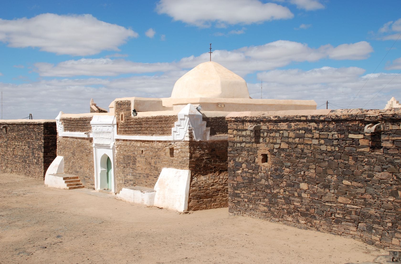A low whitewashed domed building with a green door set into long walls of rough dark stone, under a blue sky with white clouds