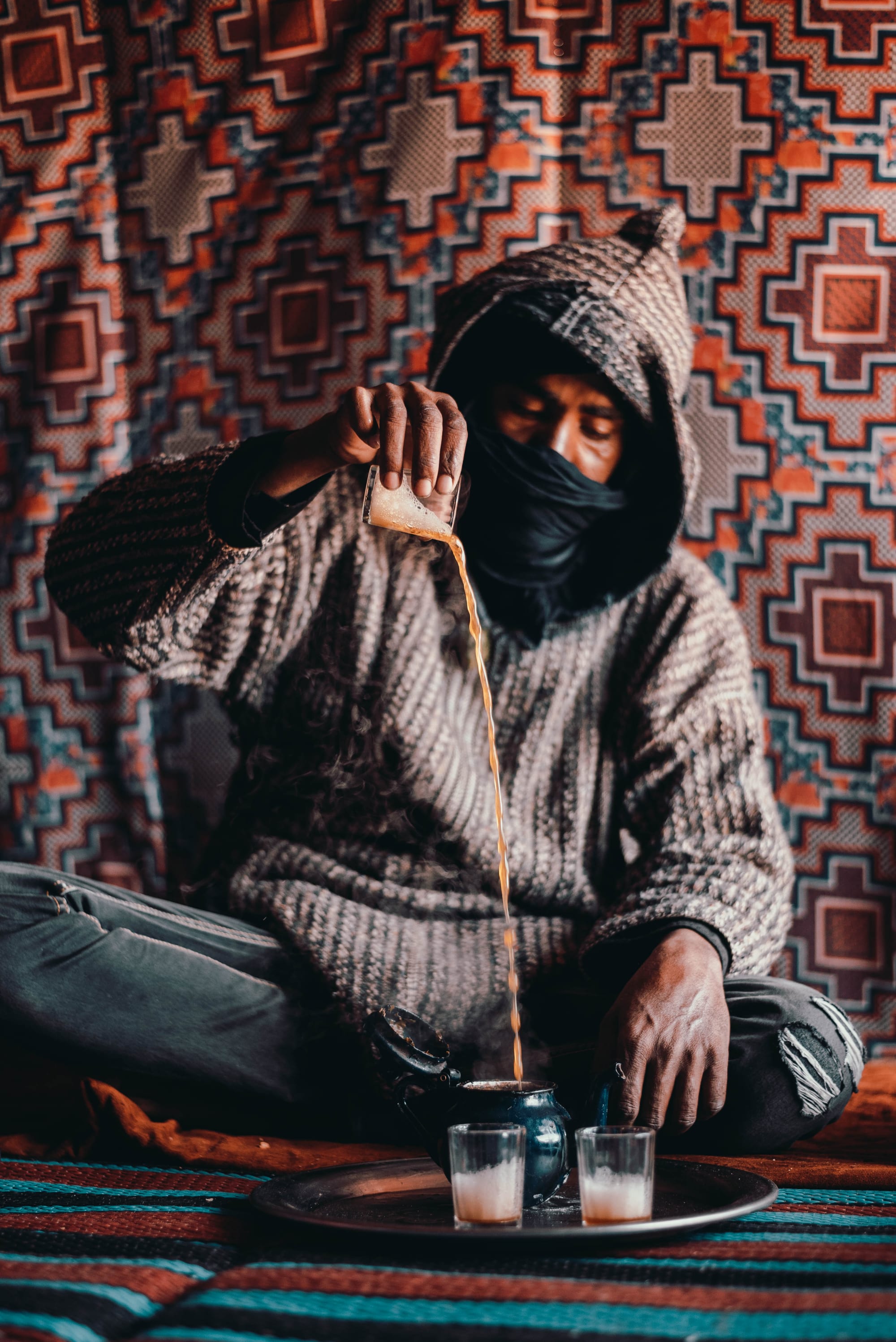 A man in a hooded knitted robe with his face wrapped in a black cloth pours tea from a small glass into a dark teapot on a round tray holding two foaming glasses, against a patterned red and orange backdrop