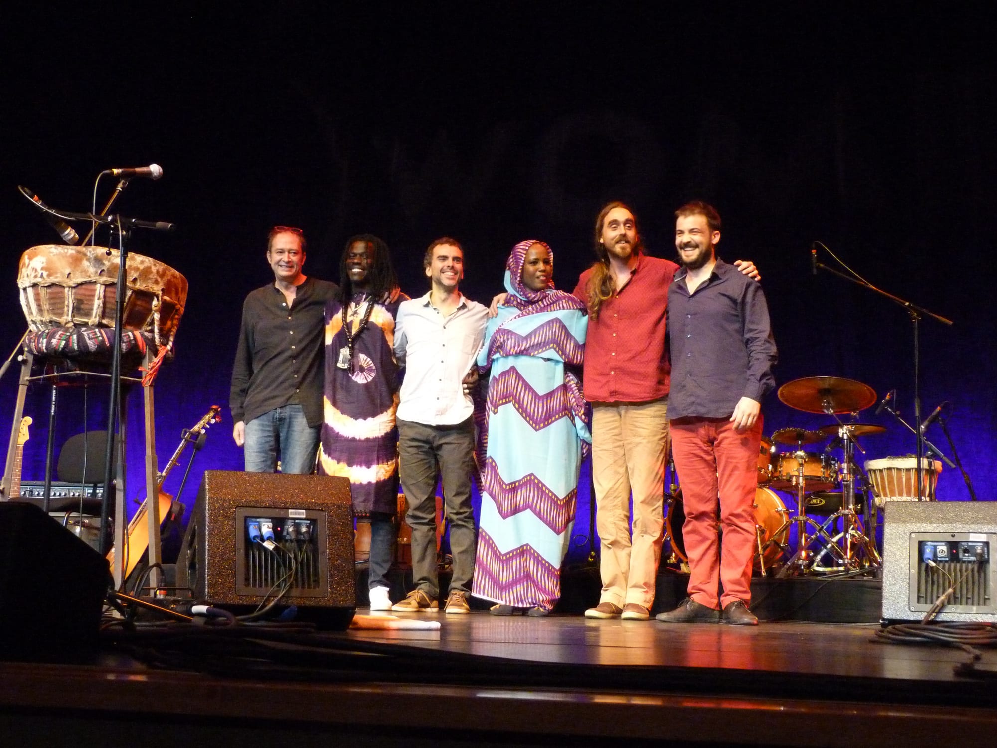 Six musicians stand arm in arm on a stage in front of drums, guitars and amplifiers, with a woman in a patterned blue and pink melhfa at the centre