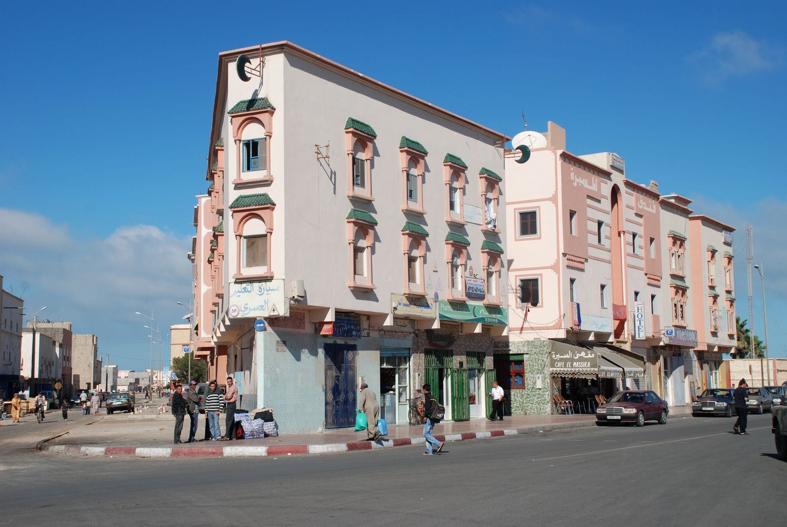 A street corner in Dakhla with pink and cream buildings, a café sign reading Cafe El Massira, and people standing and walking by