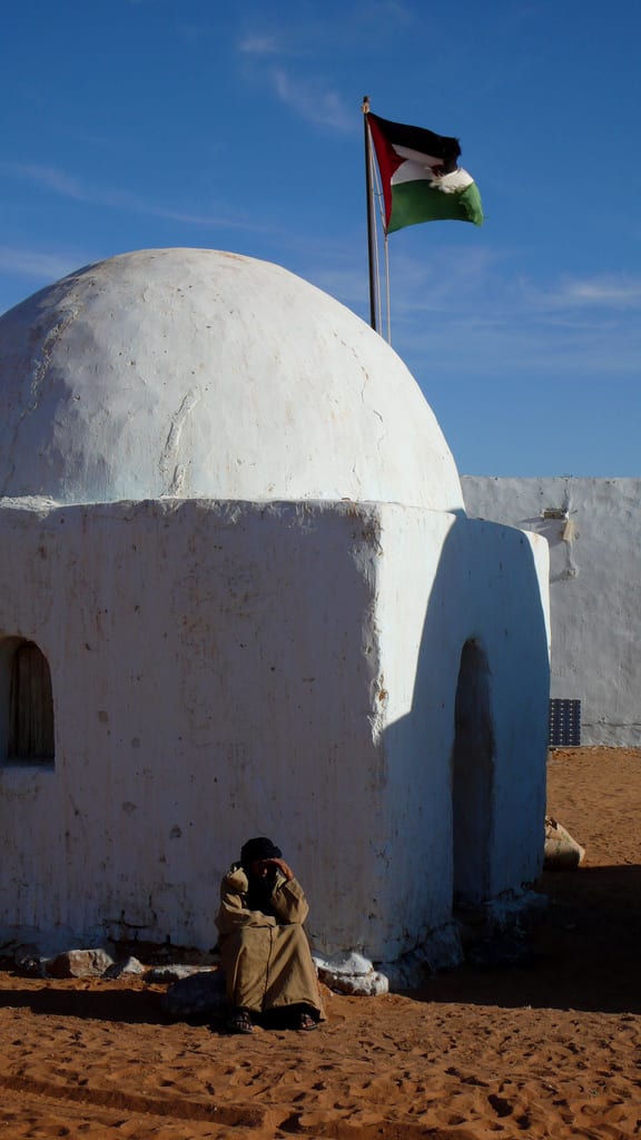 A whitewashed domed adobe dwelling in a Sahrawi refugee camp, with a person sitting in its shade and a flag flying above