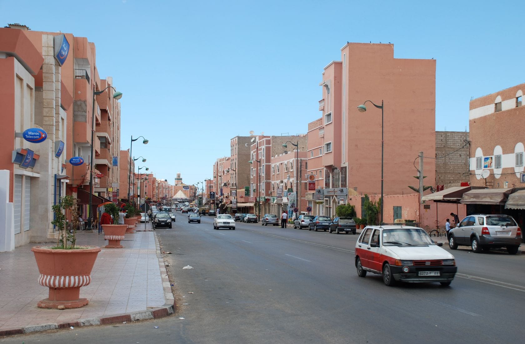 A wide, sunlit boulevard in Laayoune lined with pink and cream apartment buildings, shops and parked cars
