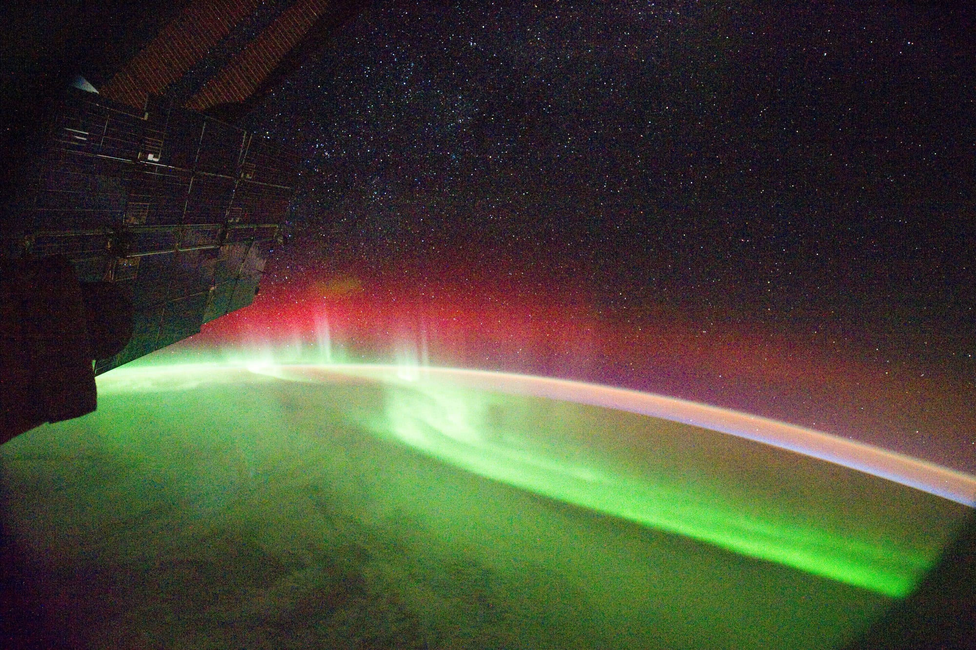 A wide band of bright green light curving along the dark Earth below, with a red glow rising above it and stars in the black sky, part of a spacecraft visible at the edge
