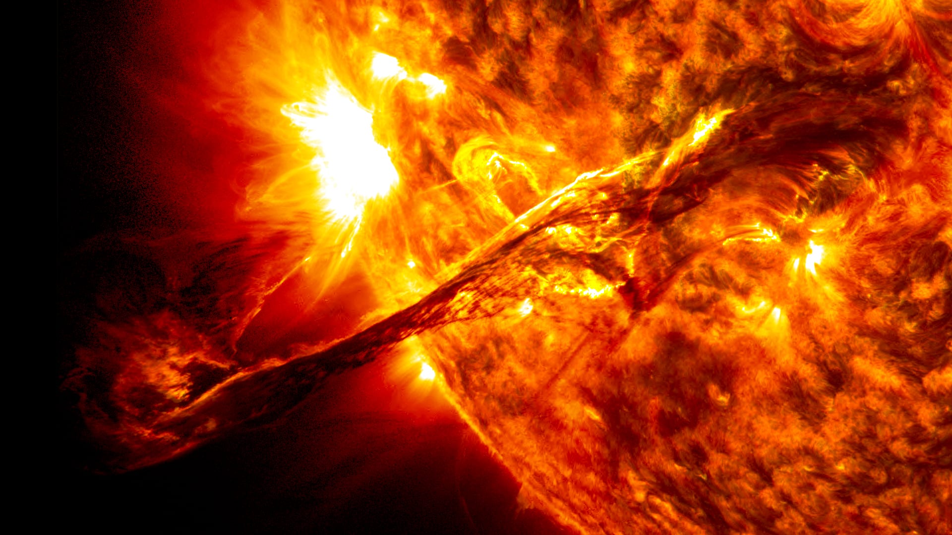 A close-up of the glowing orange surface of the Sun with a long dark twisted filament of material lifting away from it into space