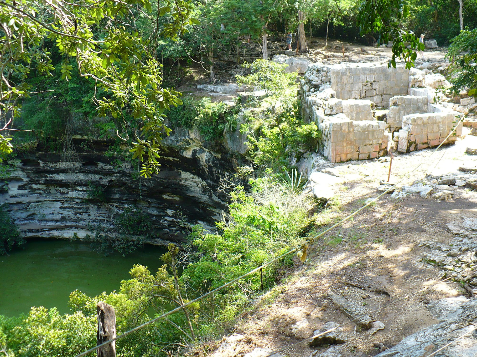 A wide sinkhole with layered limestone walls and green water below, overhung by trees, with the ruins of a small stone building on its rim