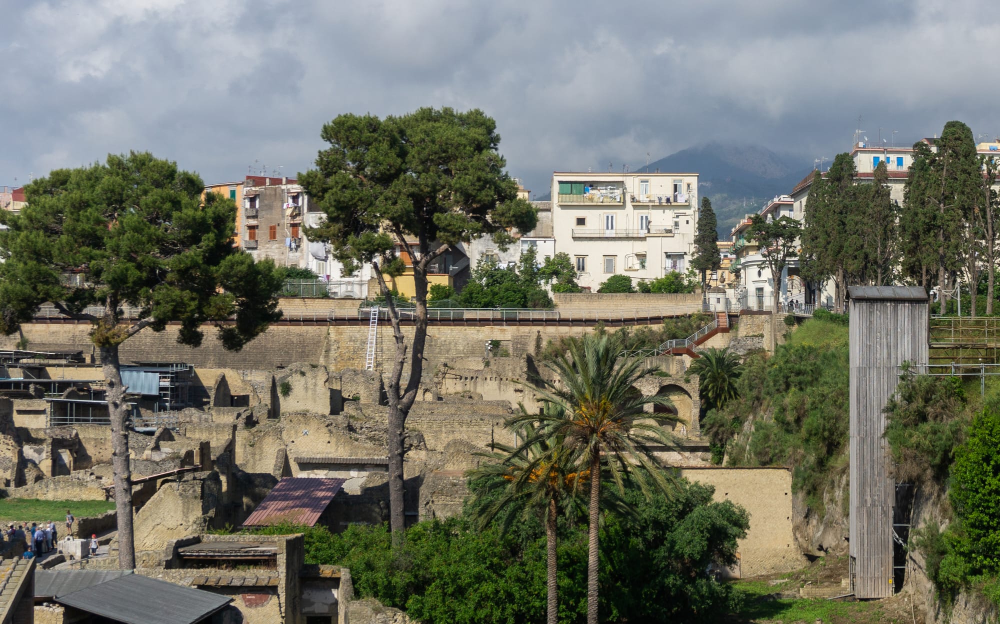 Excavated Roman ruins at the bottom of a deep pit, with apartment blocks of the modern town on the rim above and a mountain in cloud behind