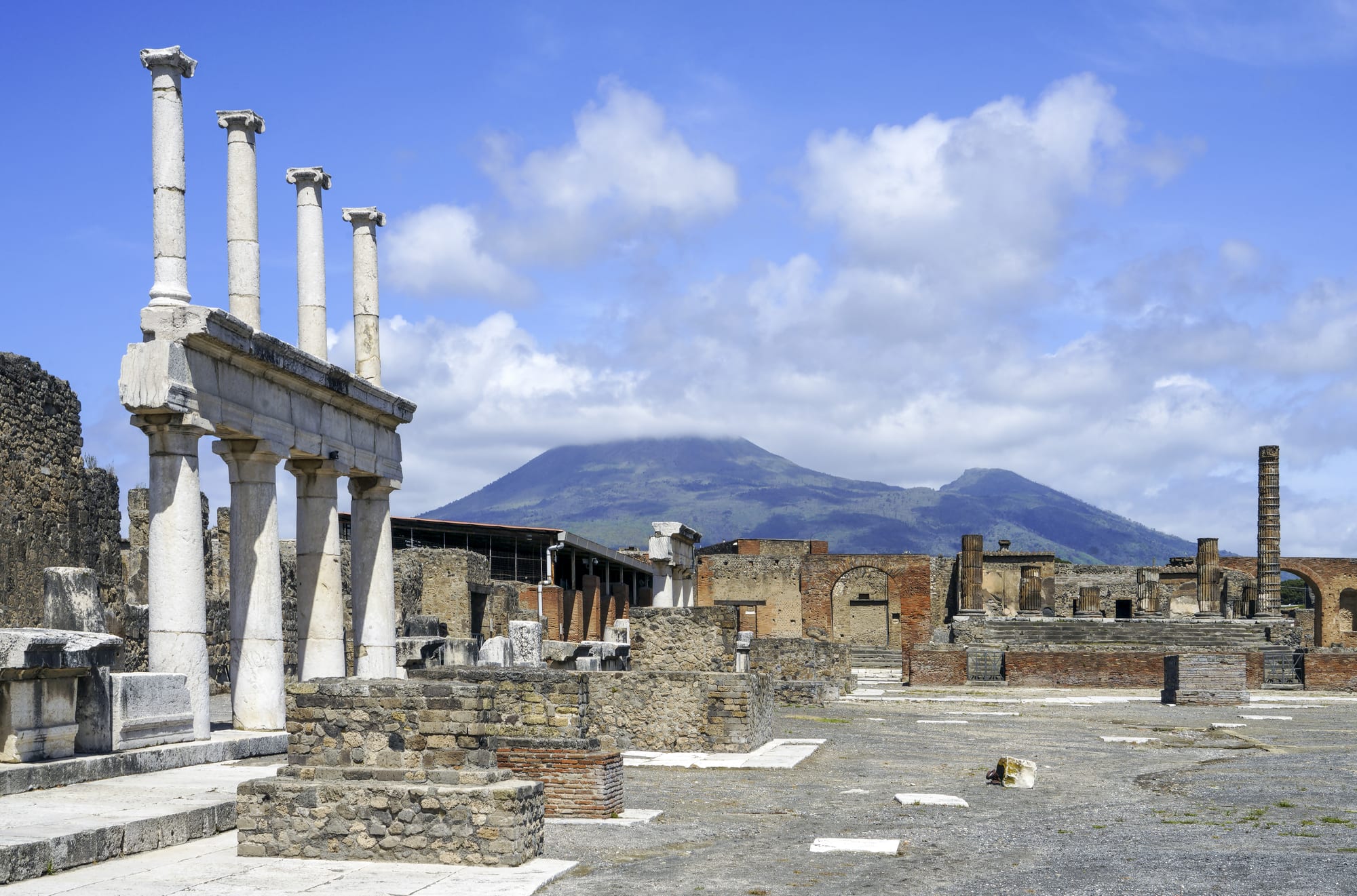 White Roman columns and the ruined walls of an ancient town square, with a broad cloud-capped volcano rising behind under a blue sky