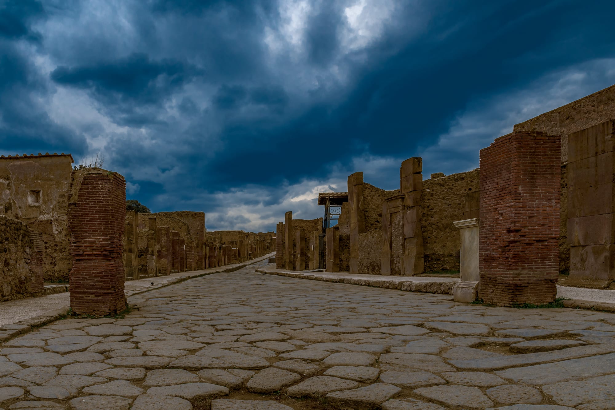A long Roman street paved with large polished stones, lined with ruined brick and stone shopfronts under a stormy sky