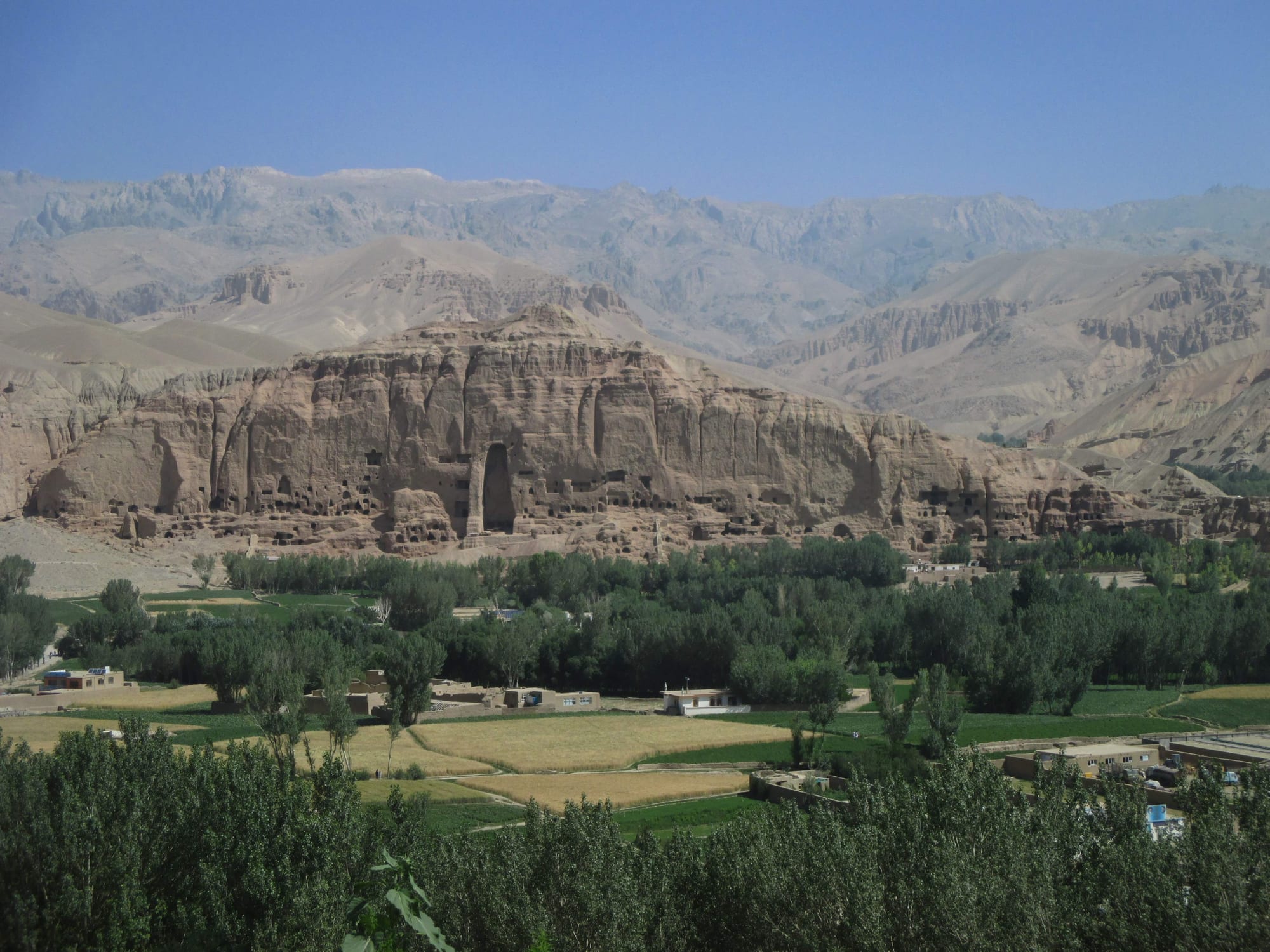 A tall, empty arched niche in a long sandstone cliff riddled with caves, above green poplar trees and fields, with mountains behind