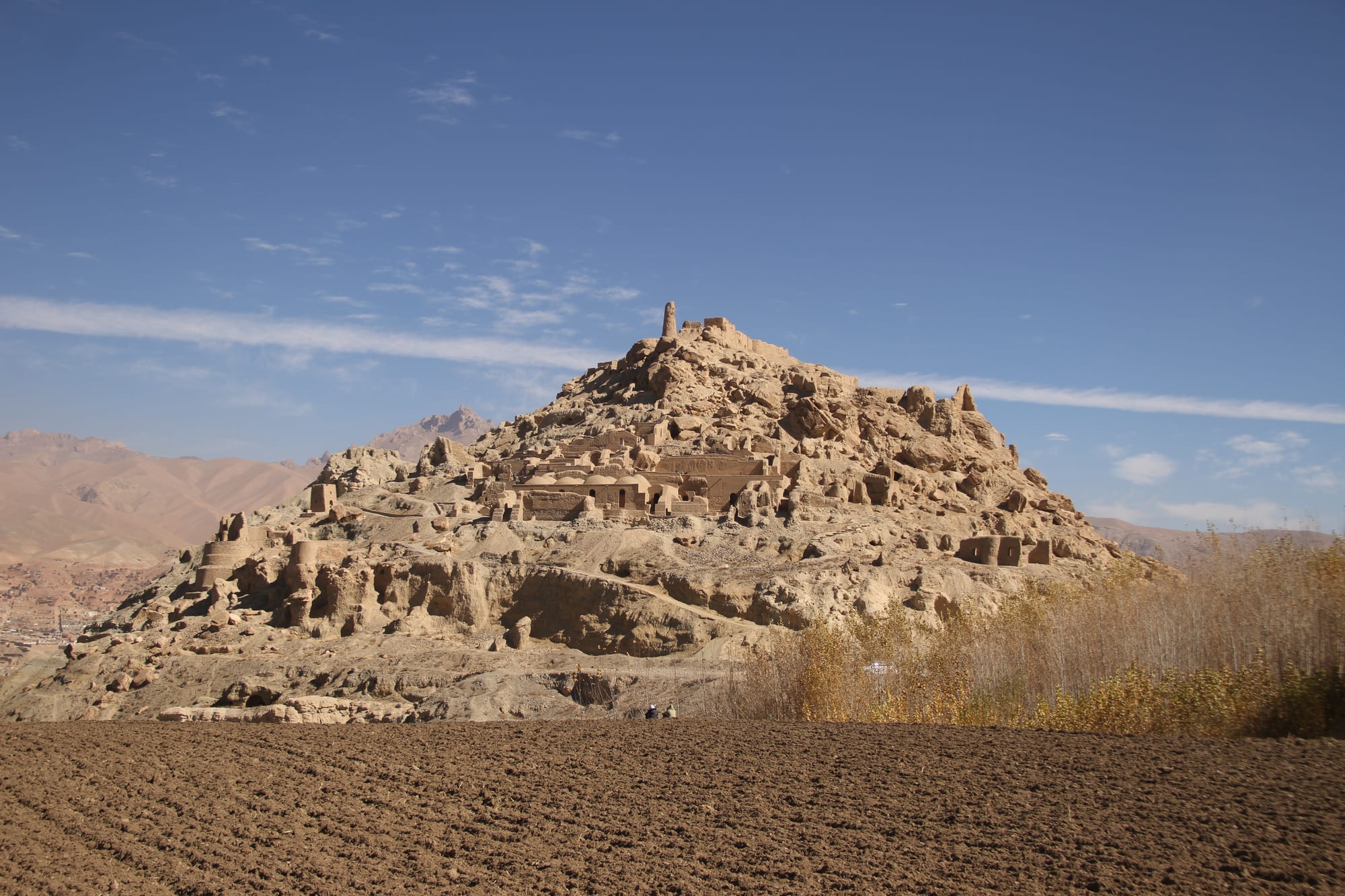 A conical hill covered with crumbling mud-brick walls and towers, rising behind a ploughed field under a blue sky