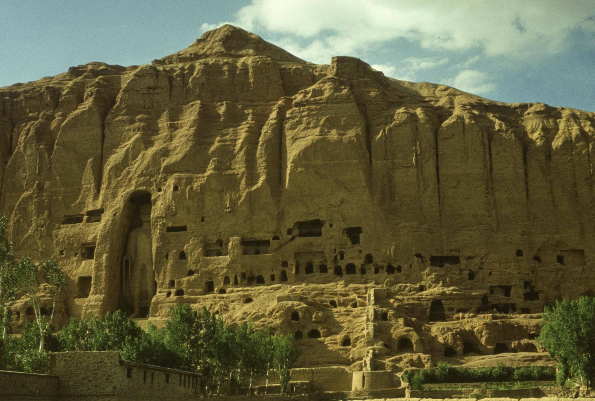 A standing Buddha figure inside a tall arched niche in a sandstone cliff dotted with cave openings, with trees below